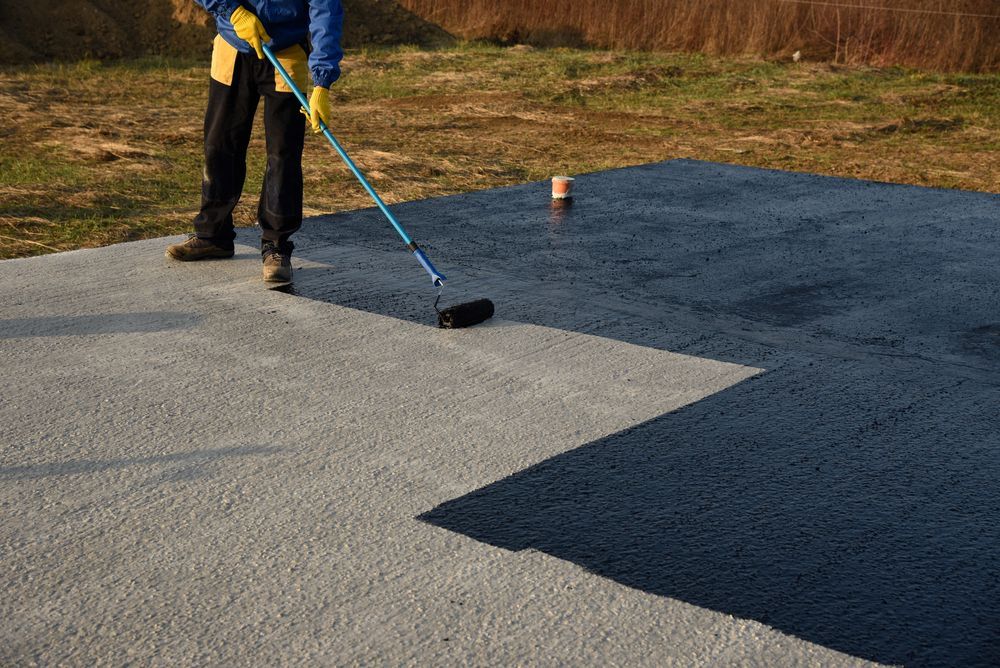Person using a roller to apply dark coating to a concrete surface outdoors.