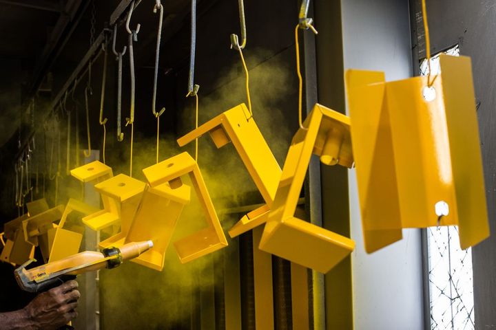 Yellow painted metal objects hanging on a conveyor belt in a workshop, being sprayed with yellow powder.