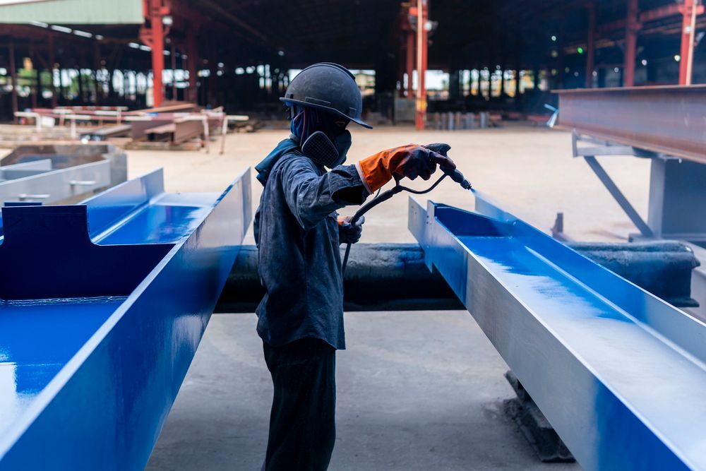 Worker spray-painting blue paint on steel beams in a factory, wearing a respirator, helmet, and gloves.