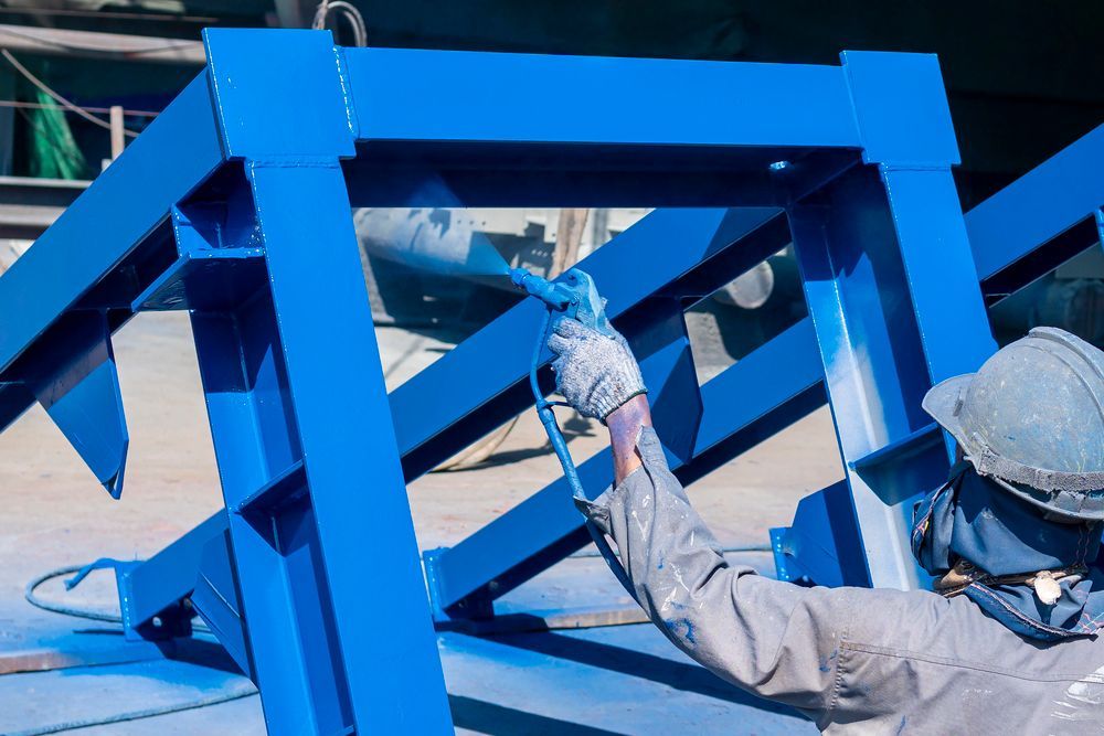 A worker in protective gear spray-paints a large blue metal structure outdoors.