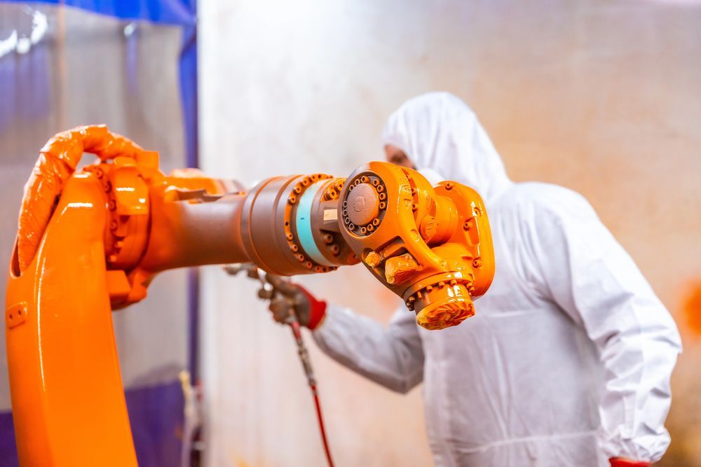 Person in protective suit spray-painting an orange robotic arm in an industrial setting.