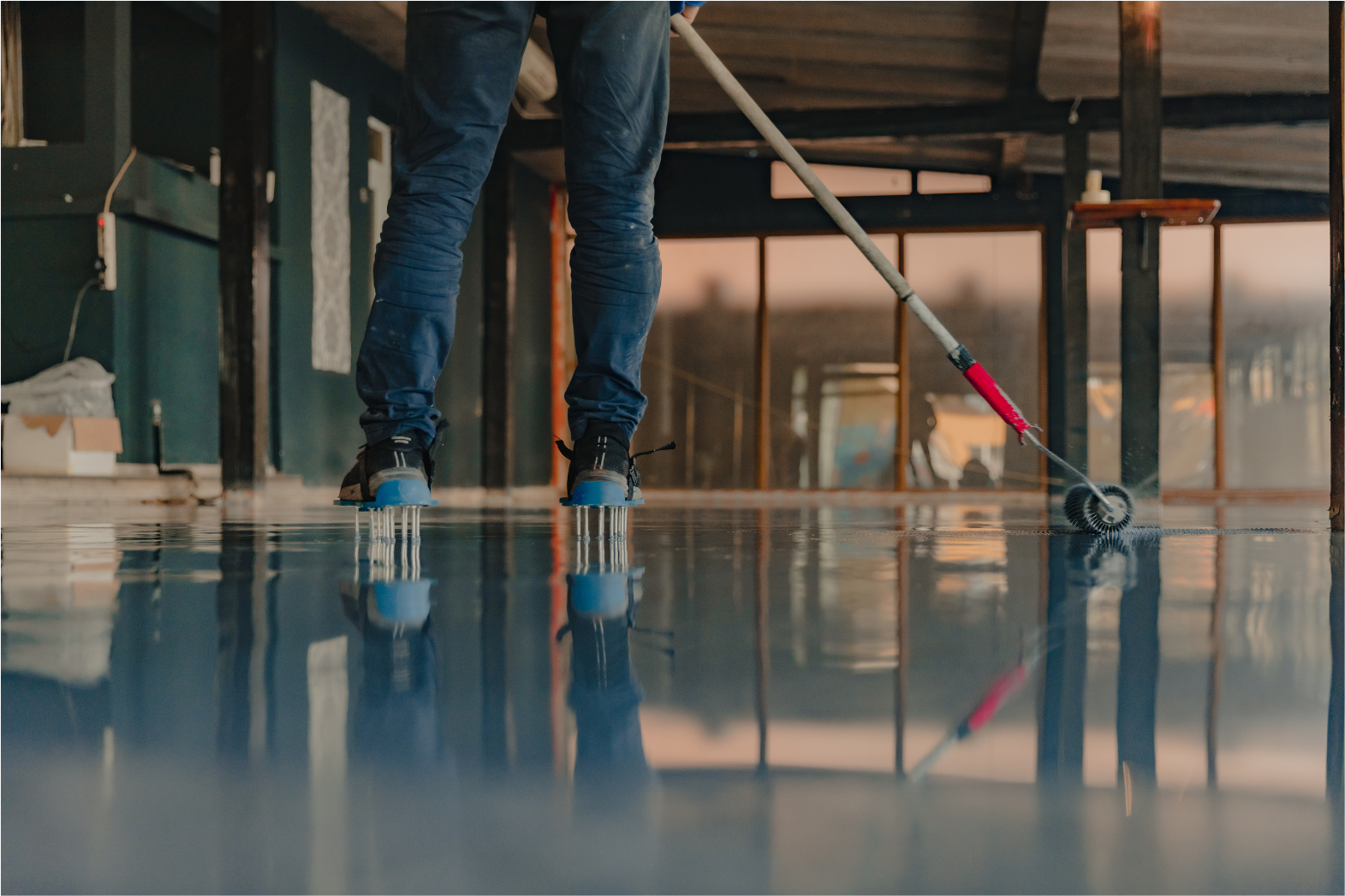 Person in jeans applying blue epoxy coating to floor with roller.