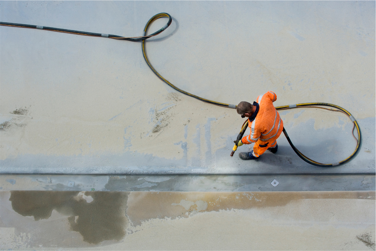 Worker in orange suit spraying concrete, long hose coiled beside him, overhead shot.