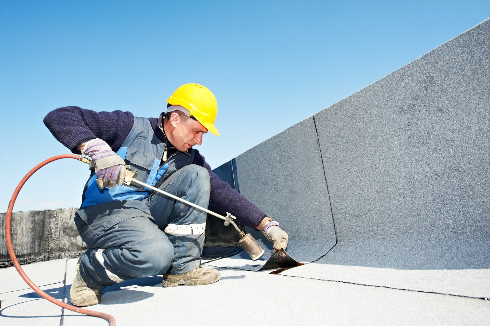 Roofer in hardhat using a torch to seal seams on a flat, black roof against a blue sky.