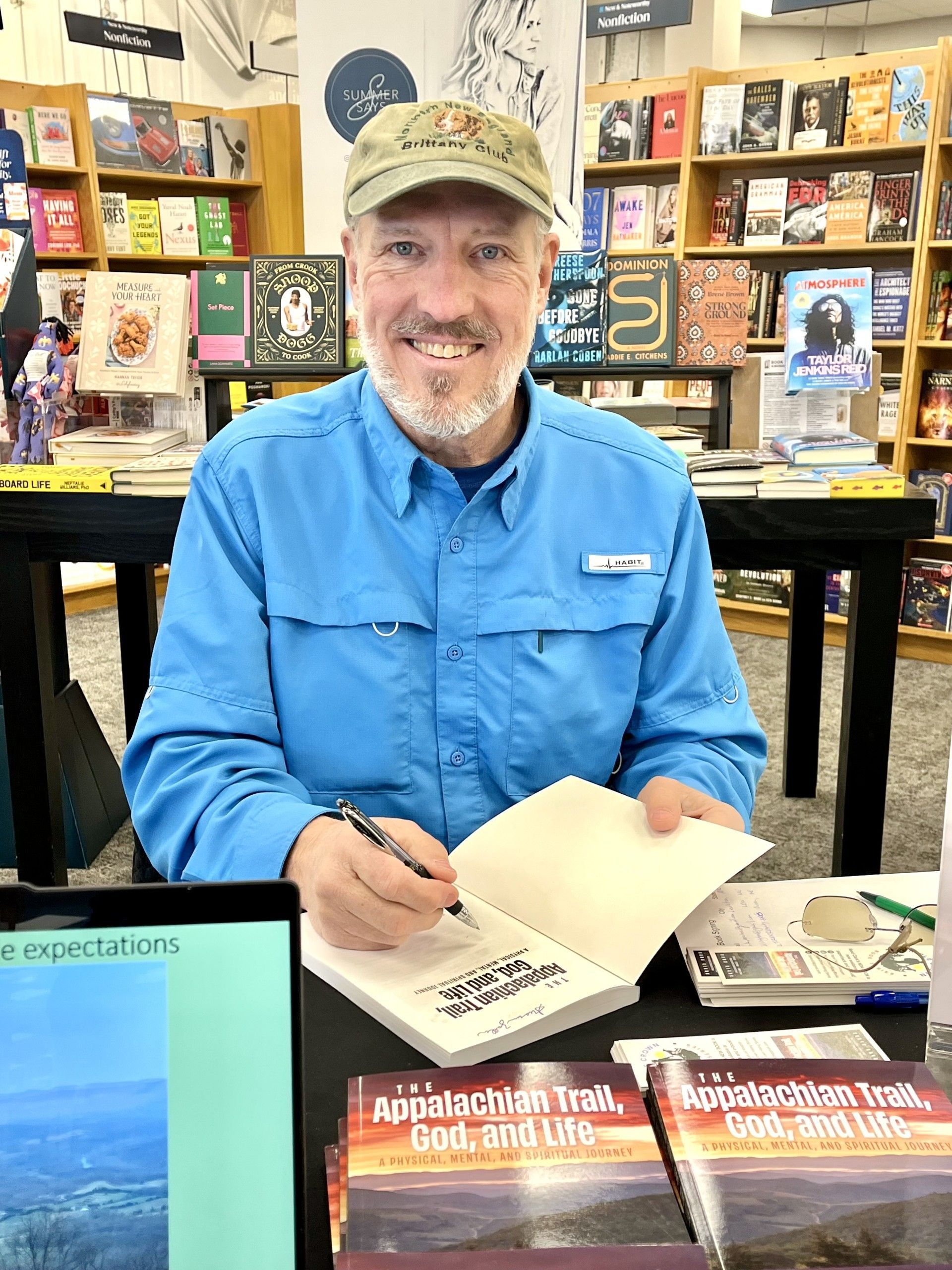 Man signing book at table in bookstore. Blue shirt, baseball cap, smiling. Bookshelves background.