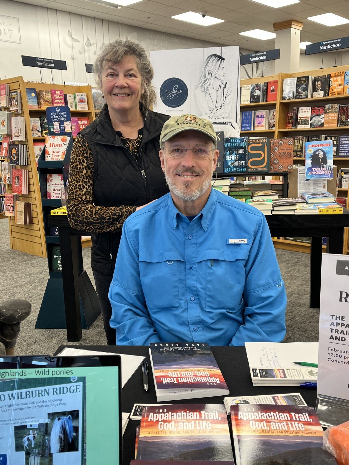 Man signing books at a table in a bookstore; woman smiles behind him; books on shelves.