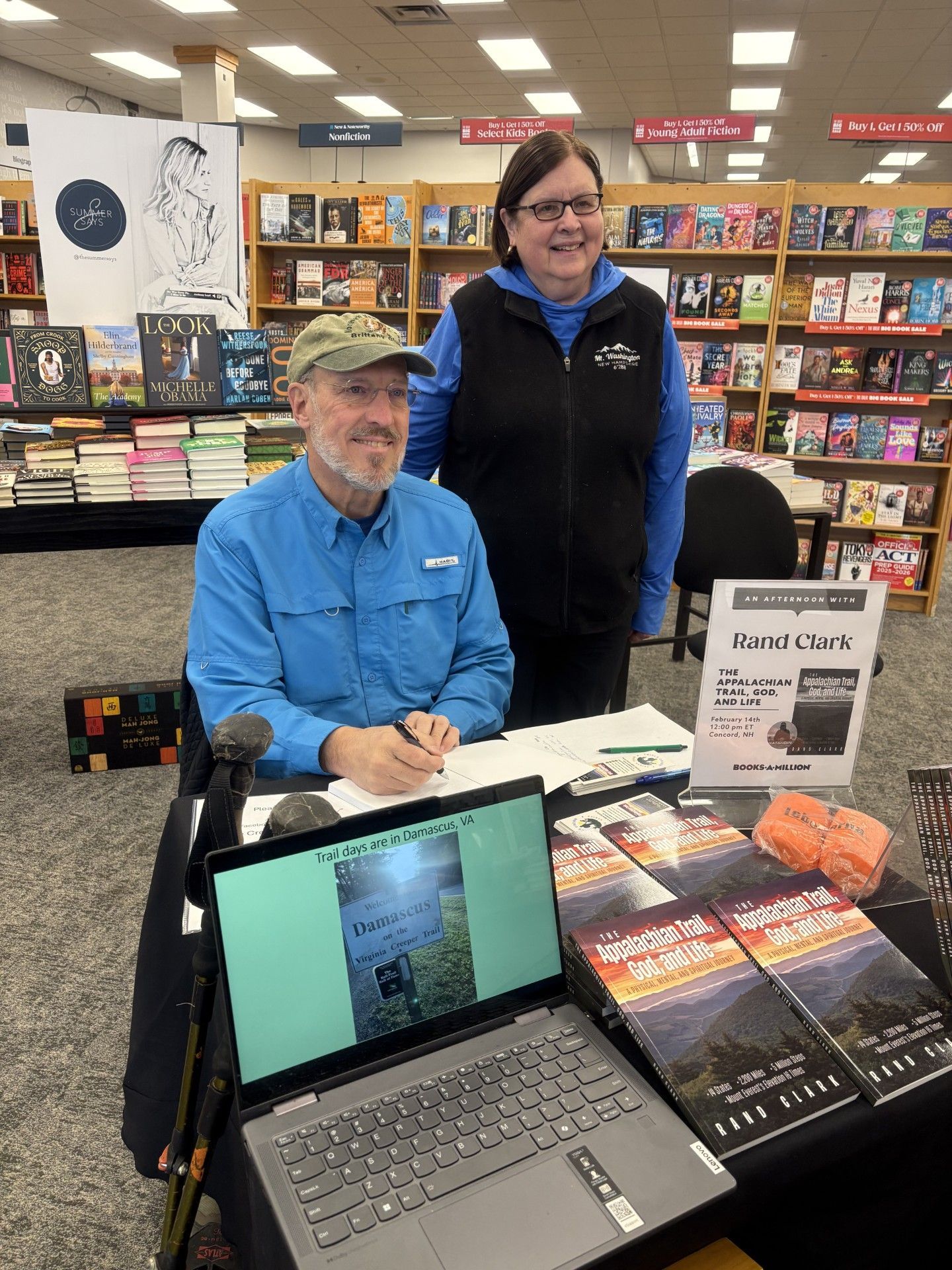 Man signing books at a table, woman standing behind. Bookstore setting with book displays.