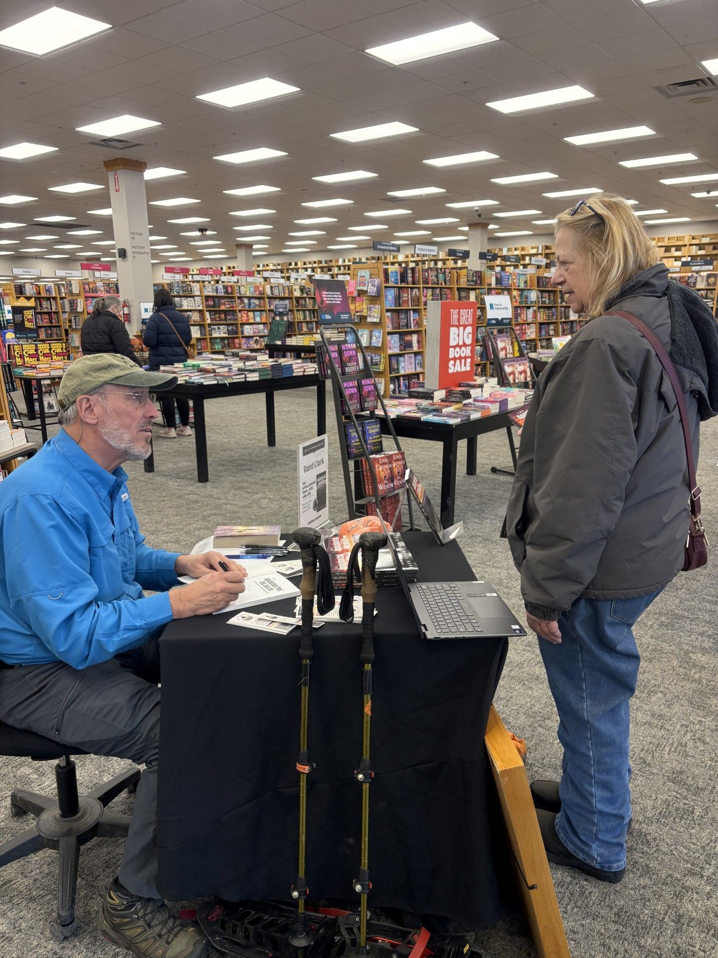 Man signing books at a table, speaking with a customer in a bookstore.