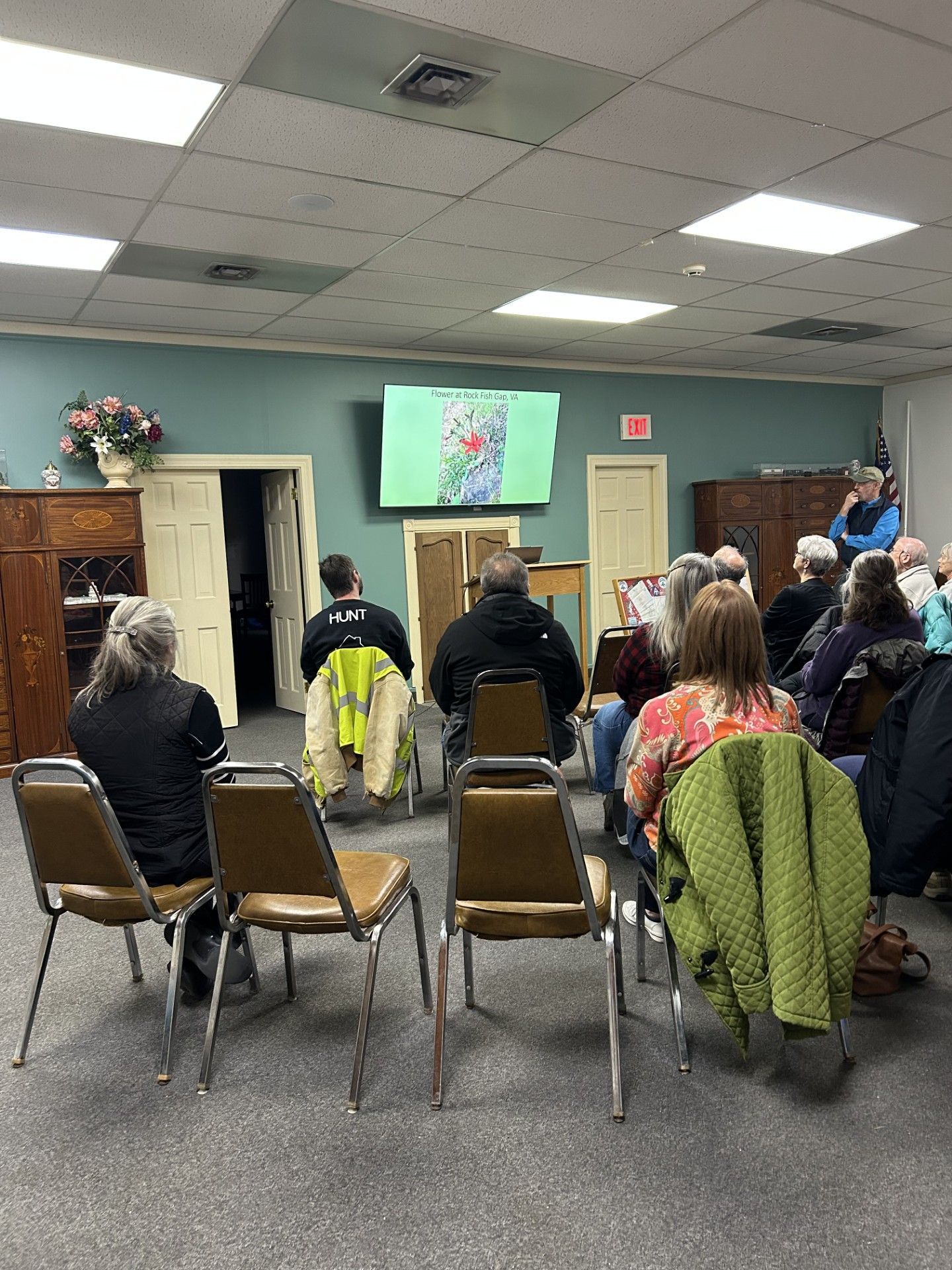 Meeting attendees watch a screen. A person speaks at front, in a room with chairs and a screen.