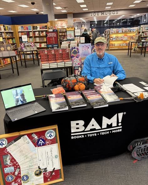 Man at a table with books for sale, possibly at a bookstore. BAM! logo on the table.