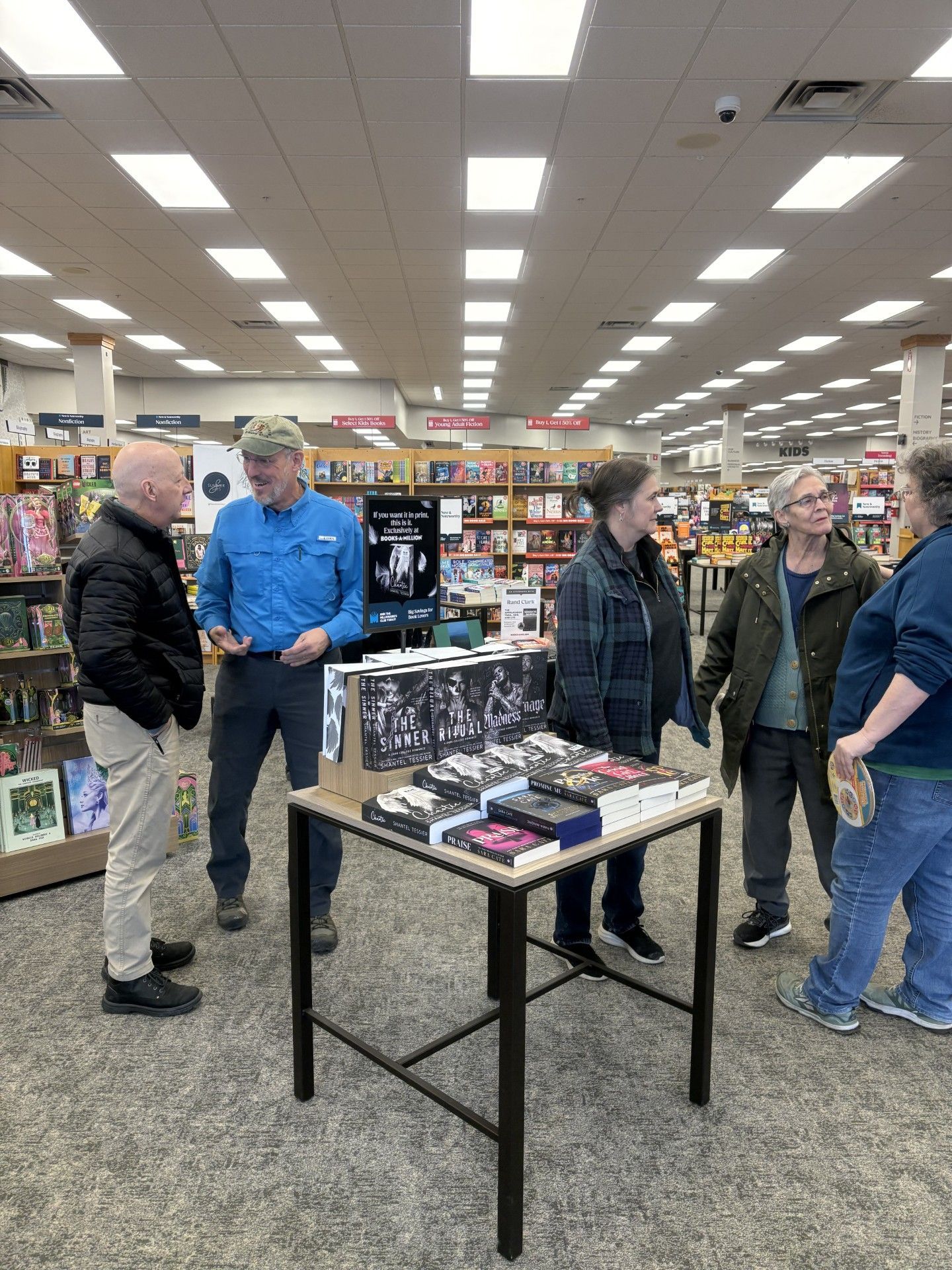 People browse books at a table inside a bookstore. Several people converse. Books are on display.