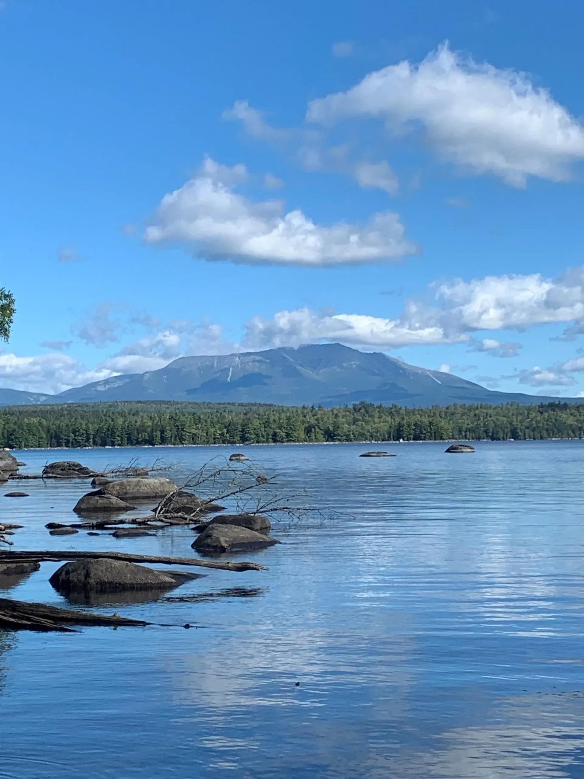 Lake scene with mountain backdrop, blue sky and clouds. Rocks in foreground, forest along shoreline.