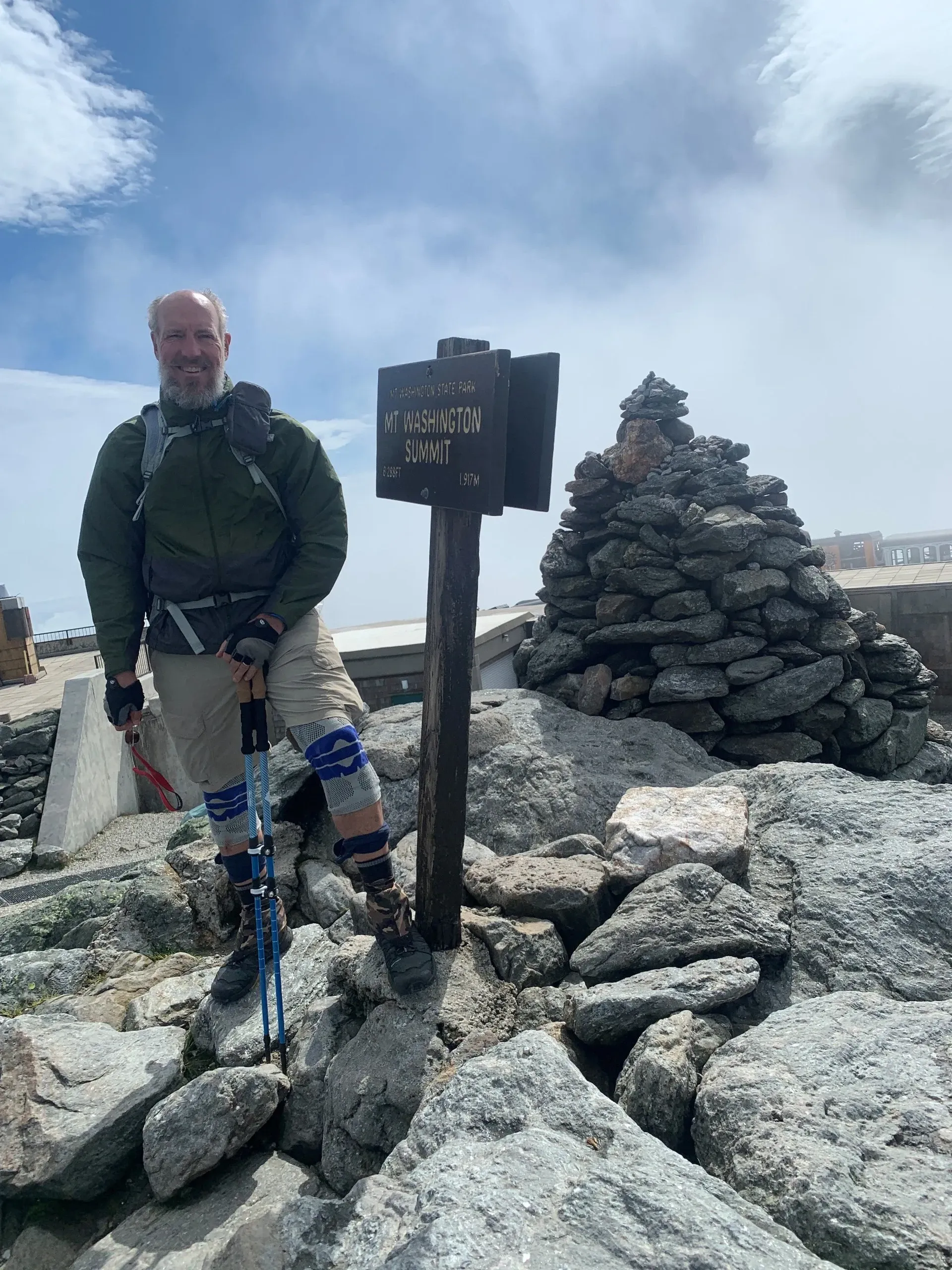 Man standing at a mountain summit sign. He wears a jacket, pack, and hiking poles, with rock cairn and cloudy sky.