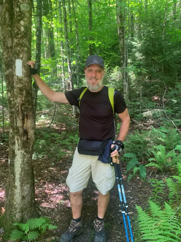 Man hiking in forest, smiling, leaning on tree with trail marker.