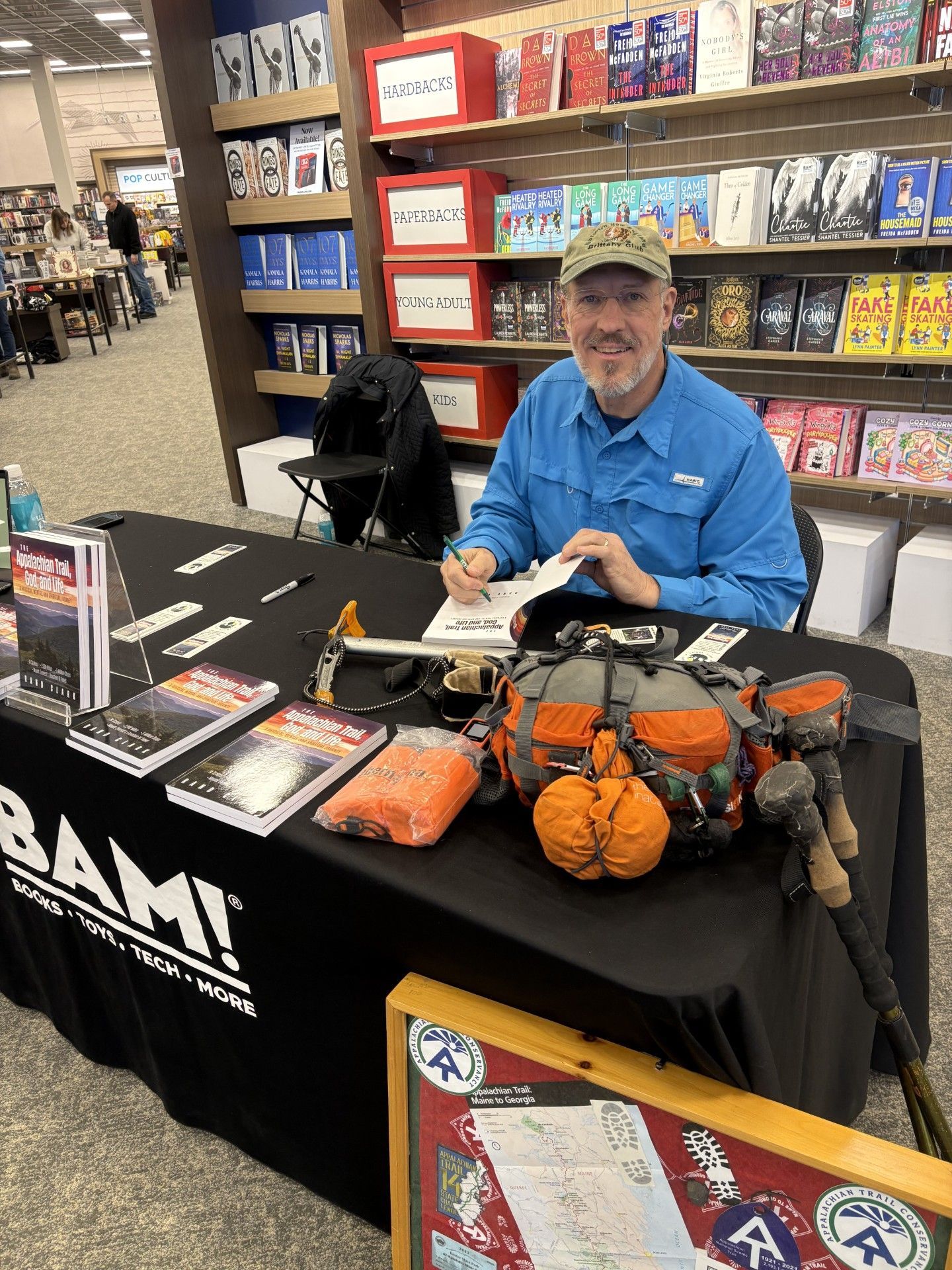Man at a table signing books, with hiking gear. Books are on the table in a bookstore setting.