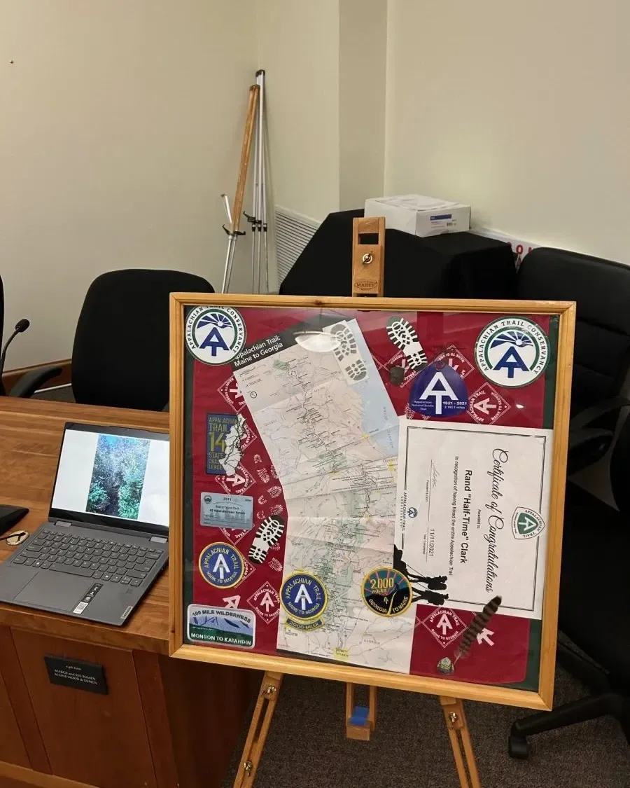 Framed Appalachian Trail memorabilia on an easel, near a laptop displaying a nature scene, in an office.
