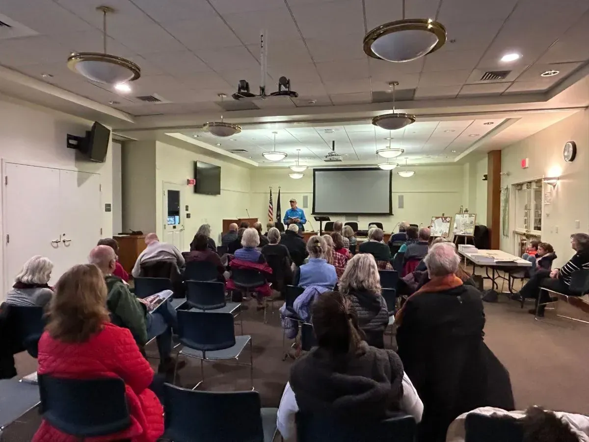 A group of people seated in a room, listening to a presentation on a screen.