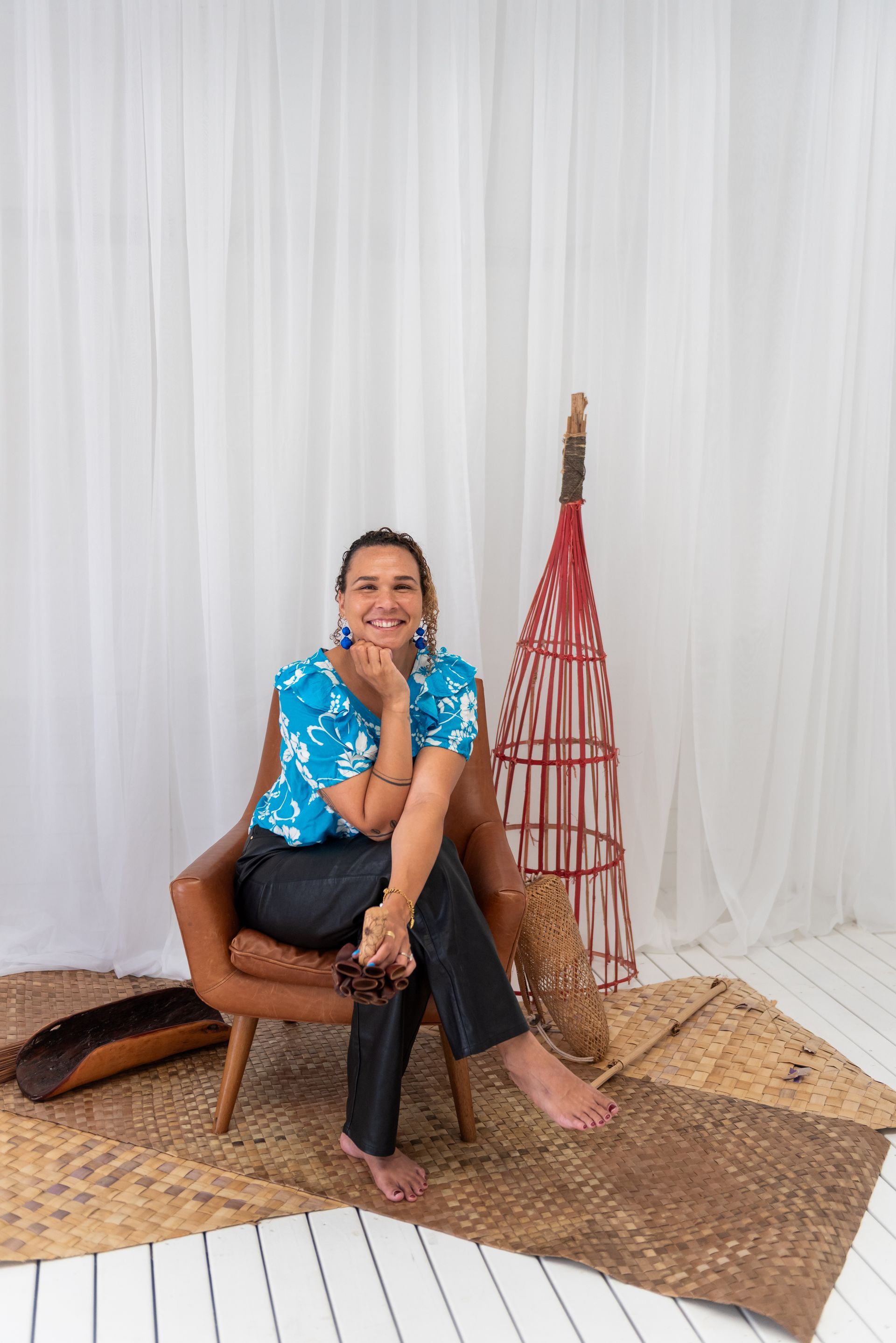 Woman sitting in chair, smiling. Wearing blue top and black pants. Indoors with decorative woven objects.