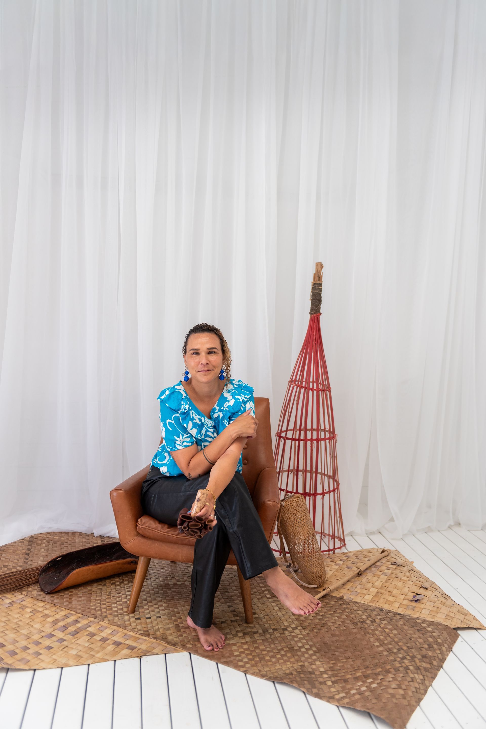 Woman sitting in a chair, wearing blue top and jeans, with woven items in a studio.