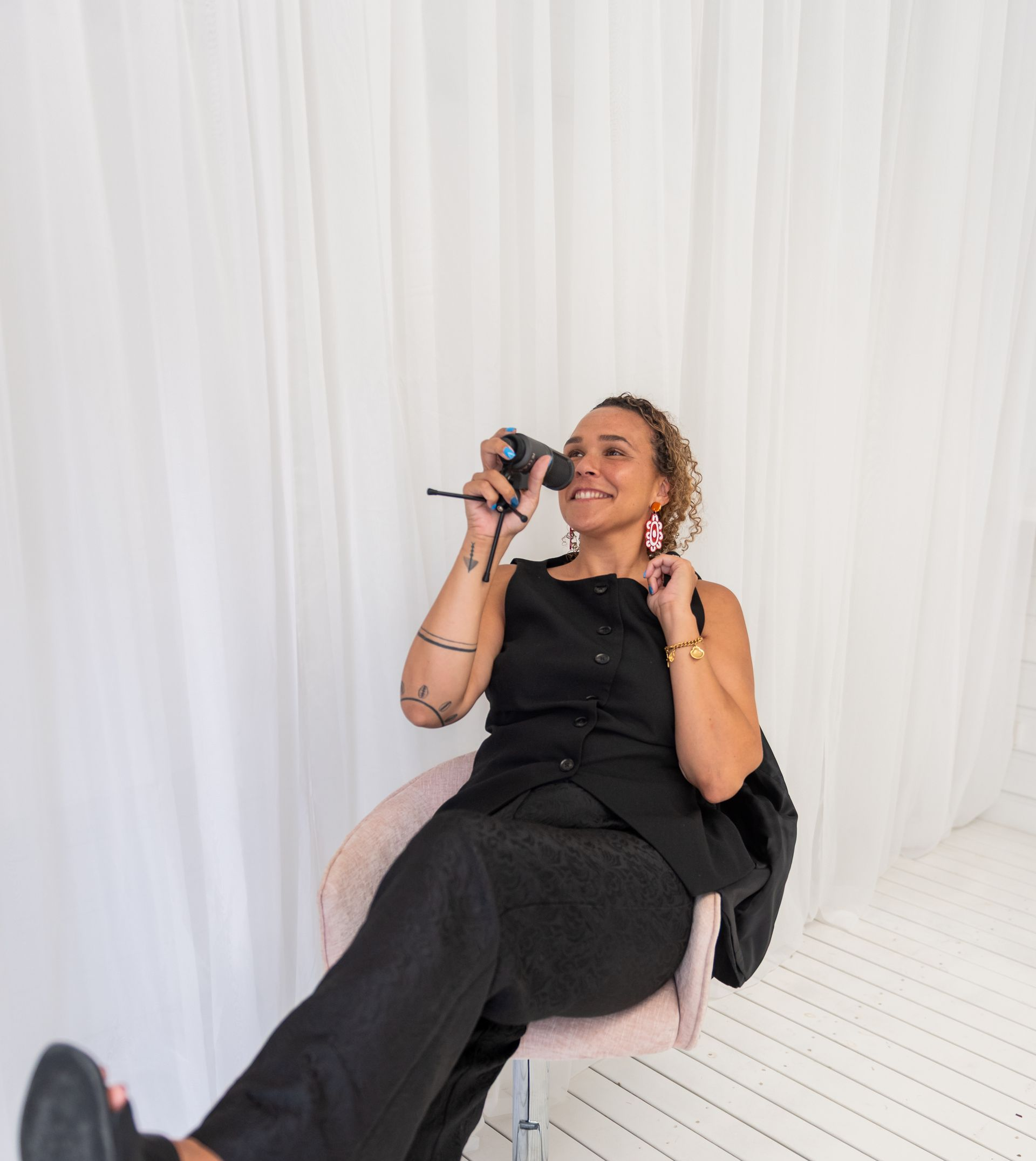 Woman in black outfit sits in a chair, holding camera, against white backdrop.