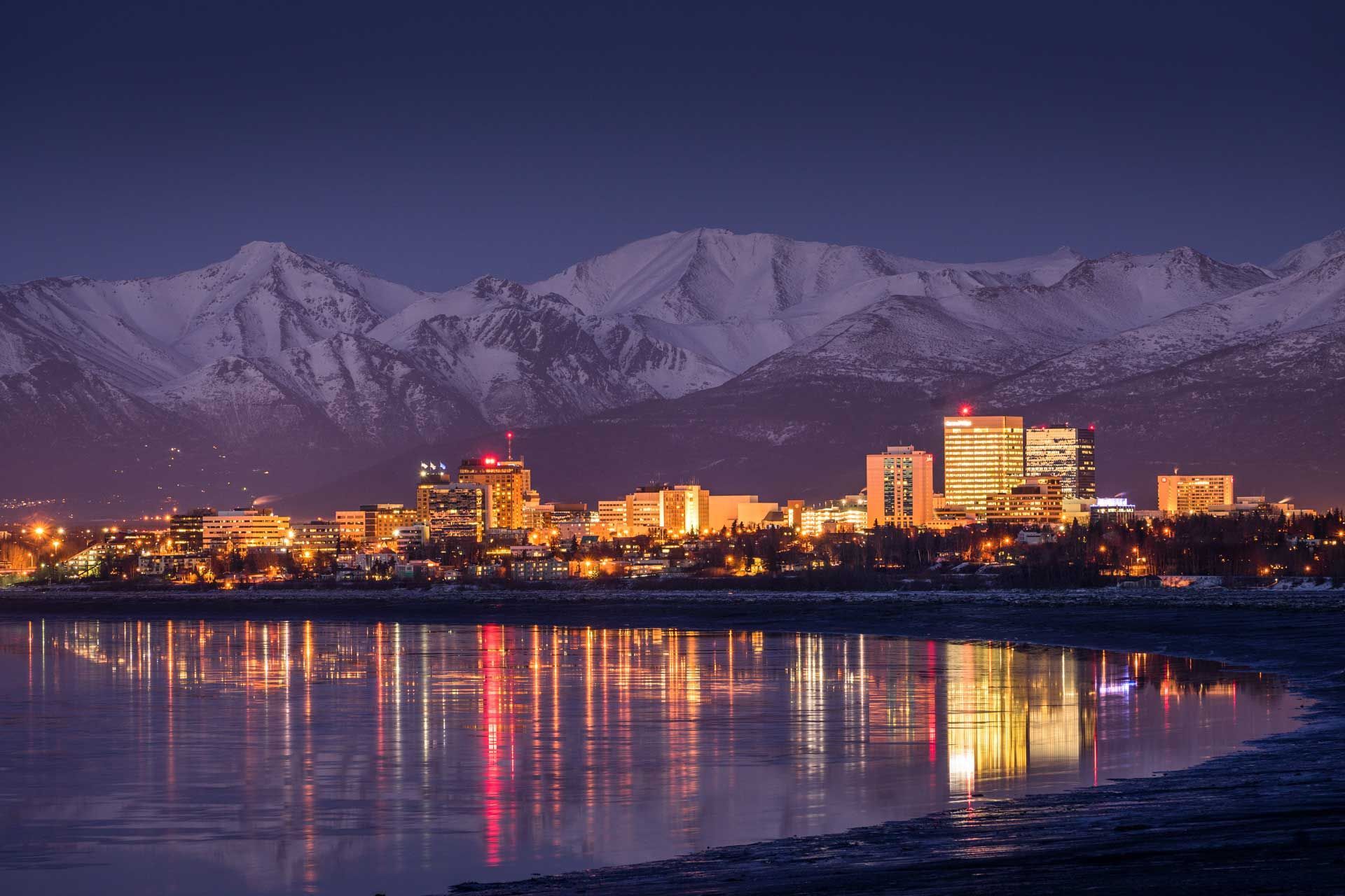 A city at night with mountains in the background and a body of water in the foreground.