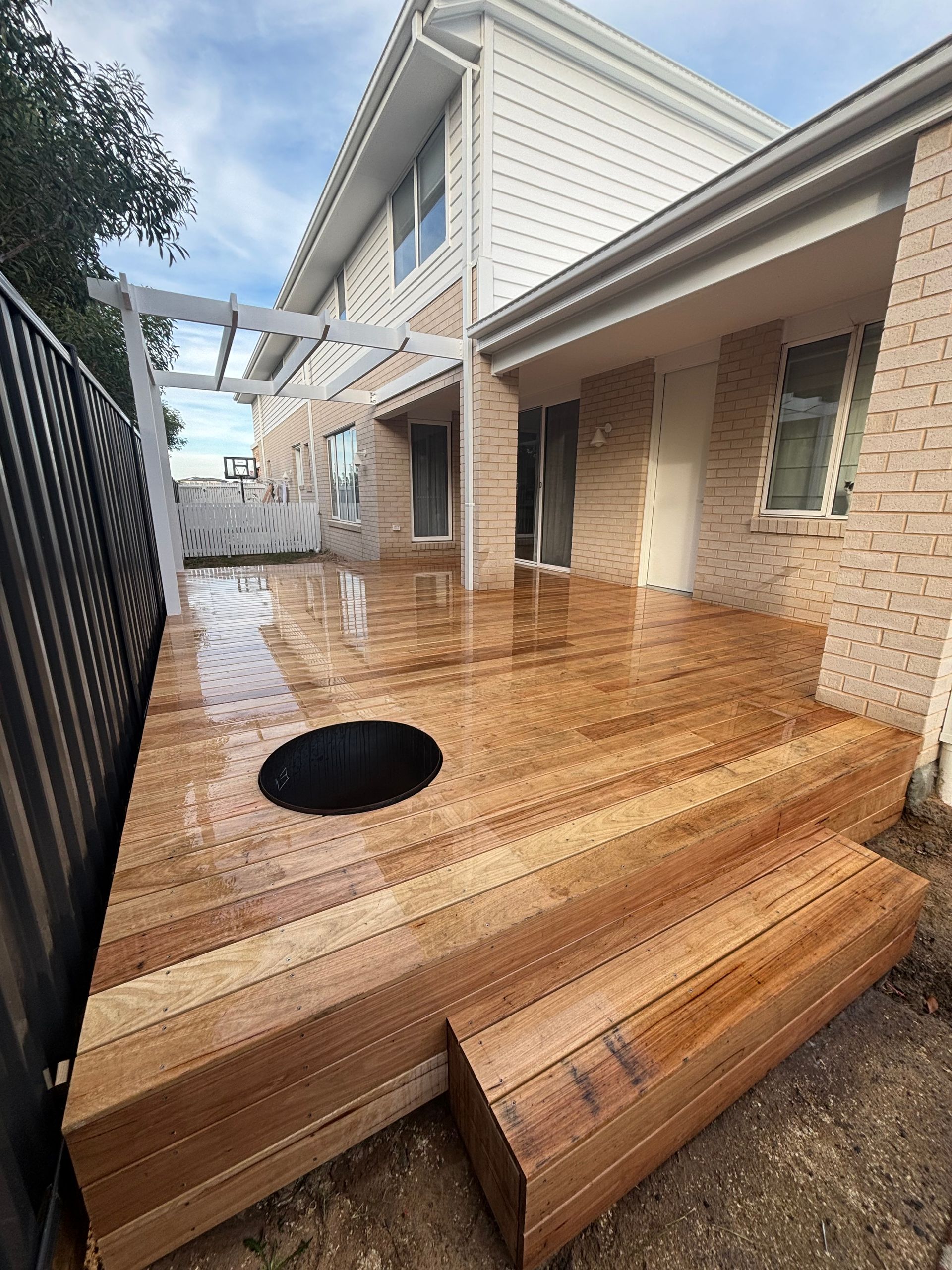 Wooden deck with built-in fire pit, next to a two-story brick house with a pergola.