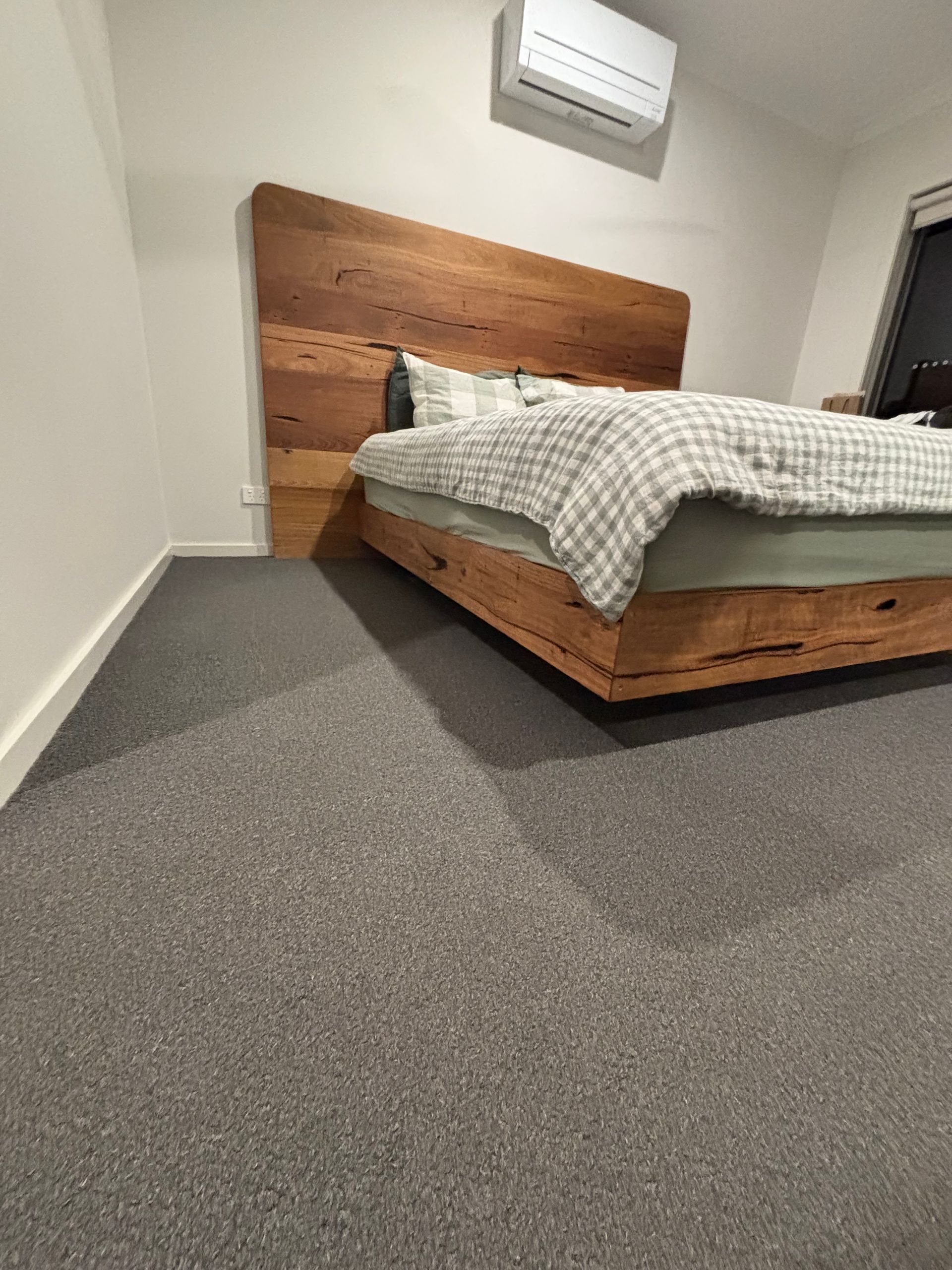 Wooden bed with headboard and bedding against a white wall; gray carpeted floor.