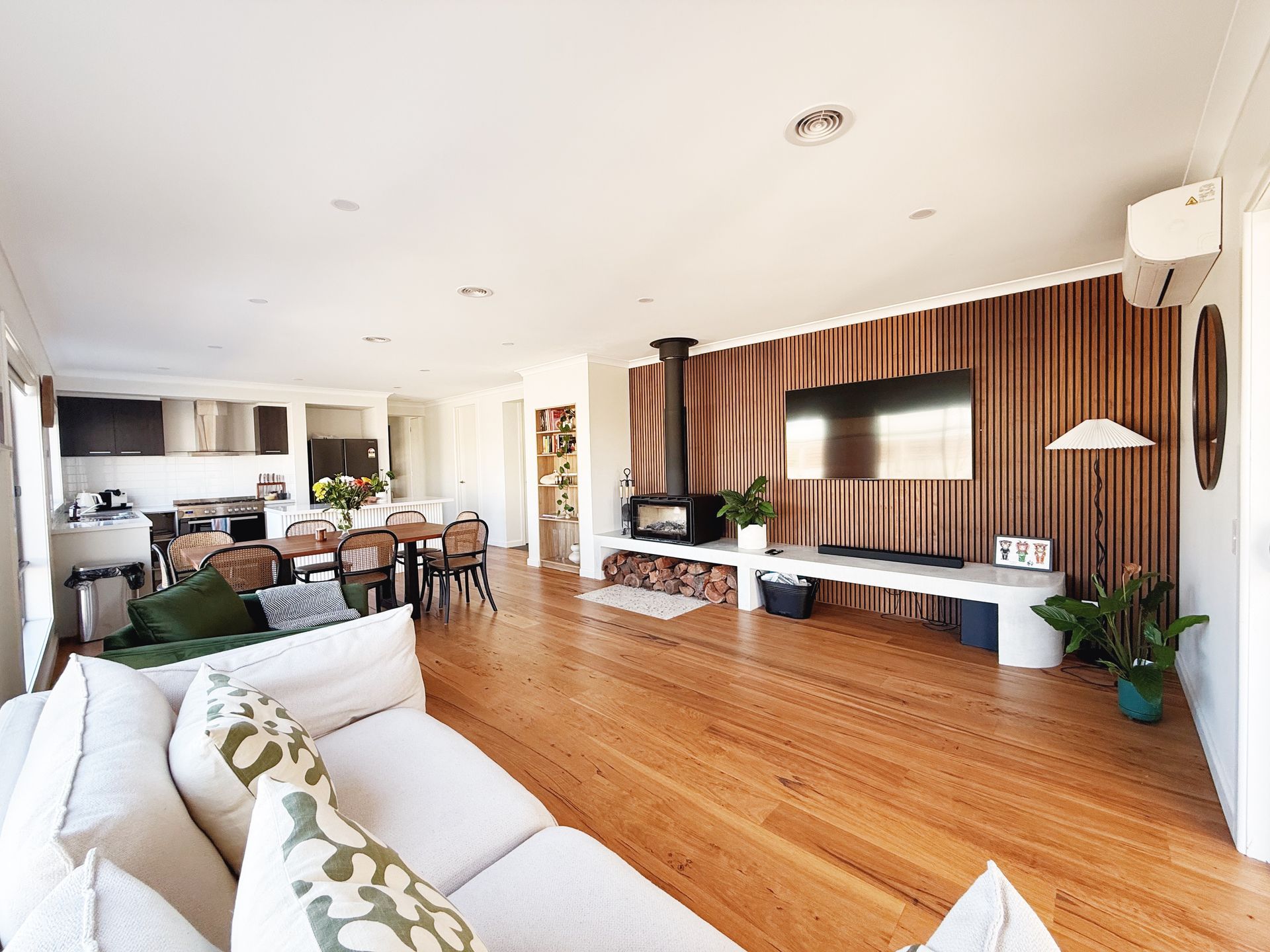 Spacious living room with wooden floors, fireplace, TV, and open kitchen. White couch with green pillows.