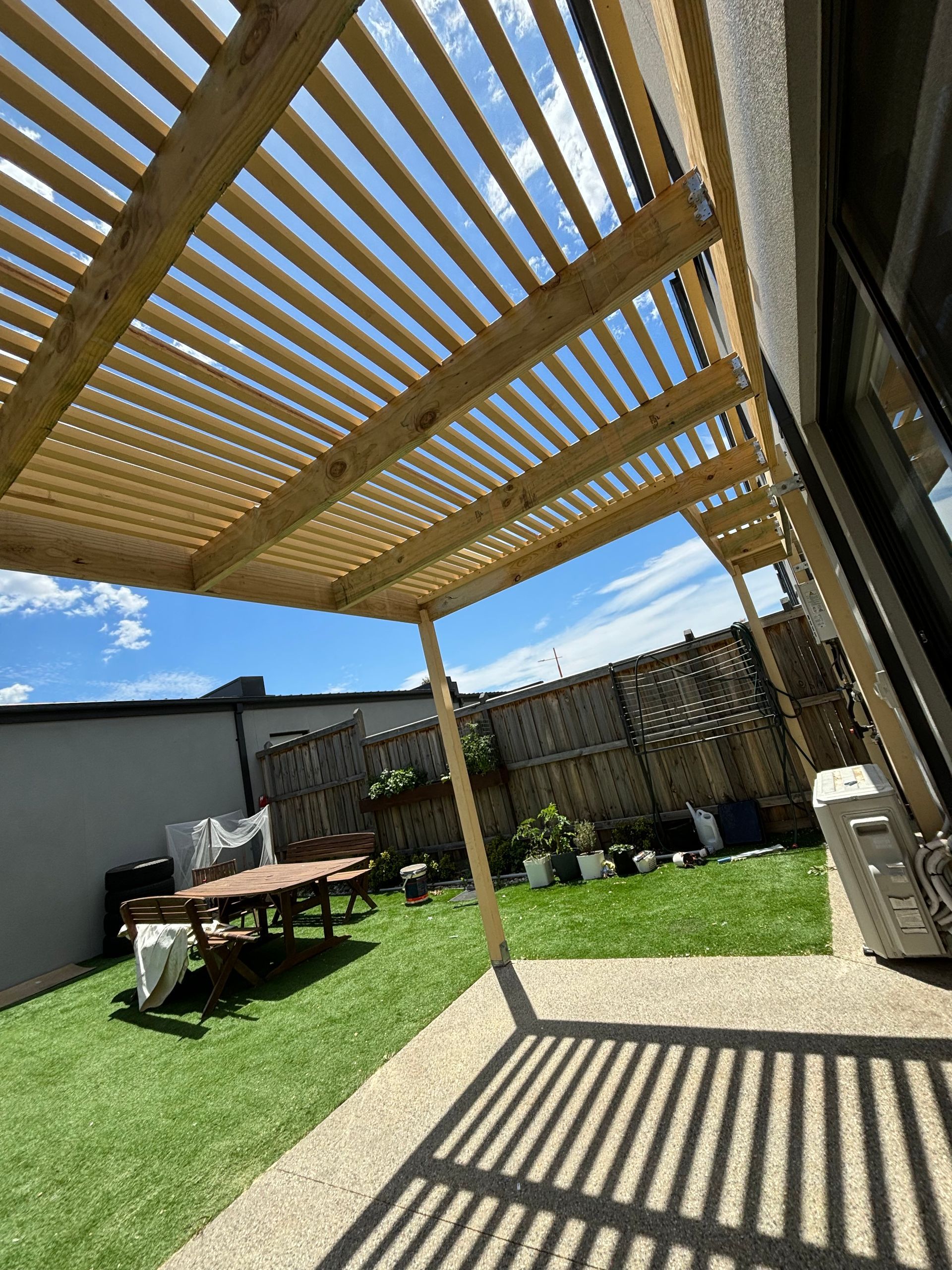 A wooden pergola in a backyard with a table and chairs underneath it.
