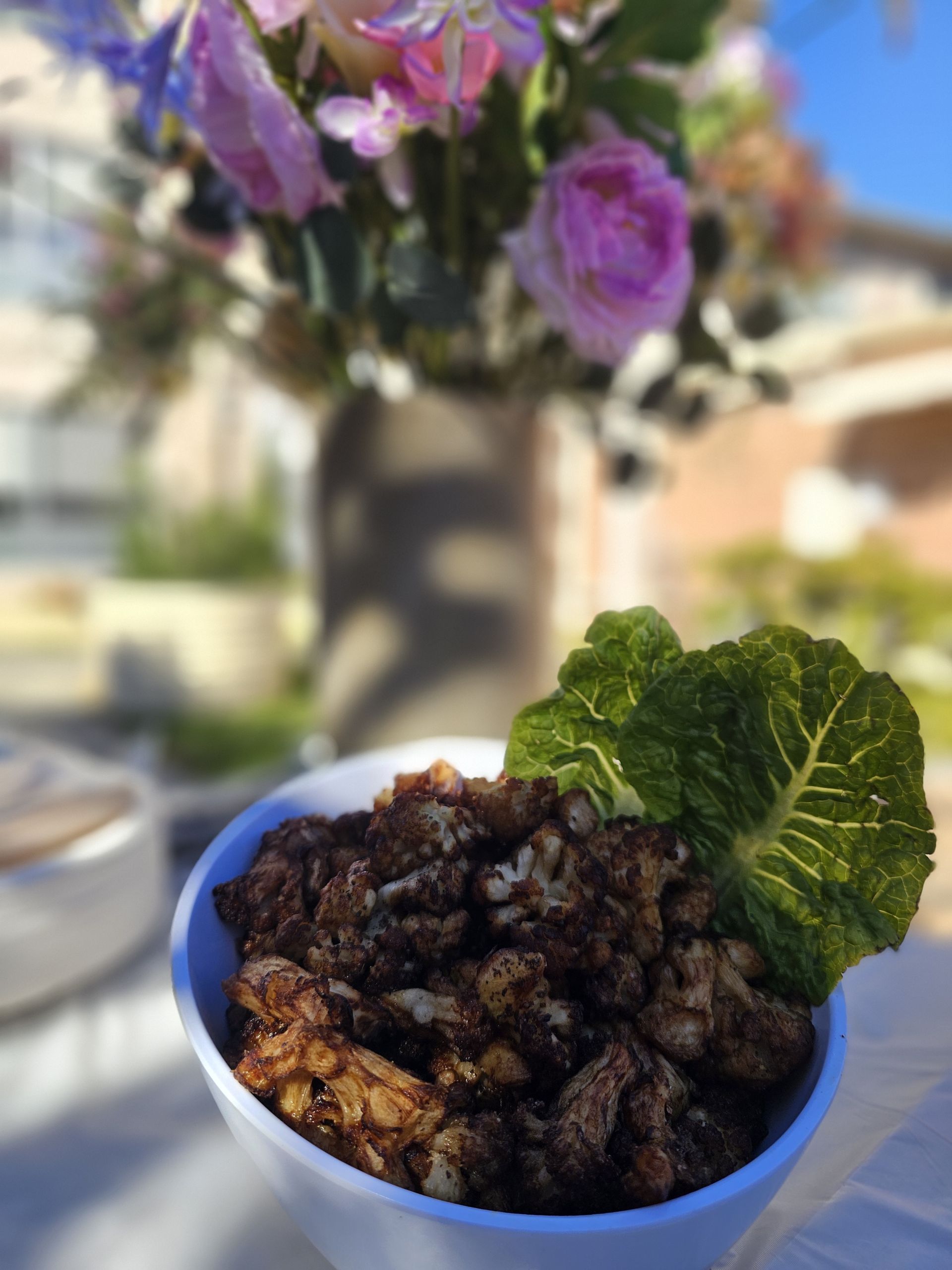 Bowl of dark-coloured food, possibly cauliflower, with a green leaf garnish. Blurred flowers in background. - The Habibiz Catering in Wingham NSW