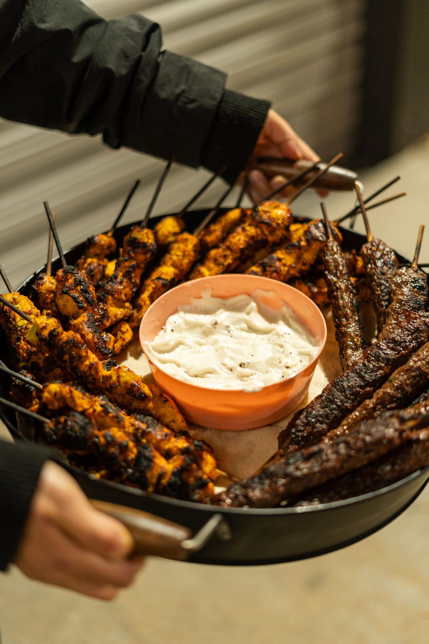 Person holding a tray of grilled kebabs with a central bowl of white sauce. — The Habibiz Catering In Wingham, NSW