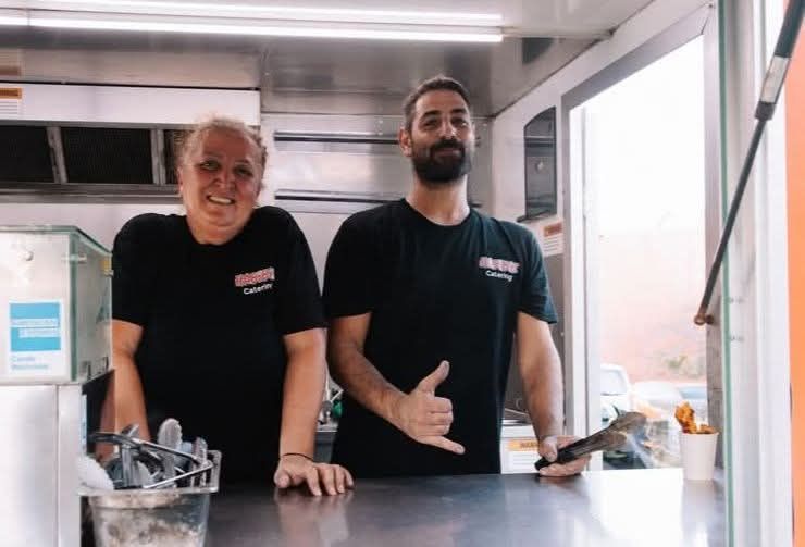 Two people in a food truck: one smiles, one gives a shaka. Both wear black shirts. Counter, open window. — The Habibiz Catering In Wingham, NSW