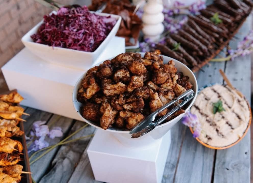 A buffet table with bowls of food like cauliflower, slaw, and hummus, on white pedestals. — The Habibiz Catering In Wingham, NSW
