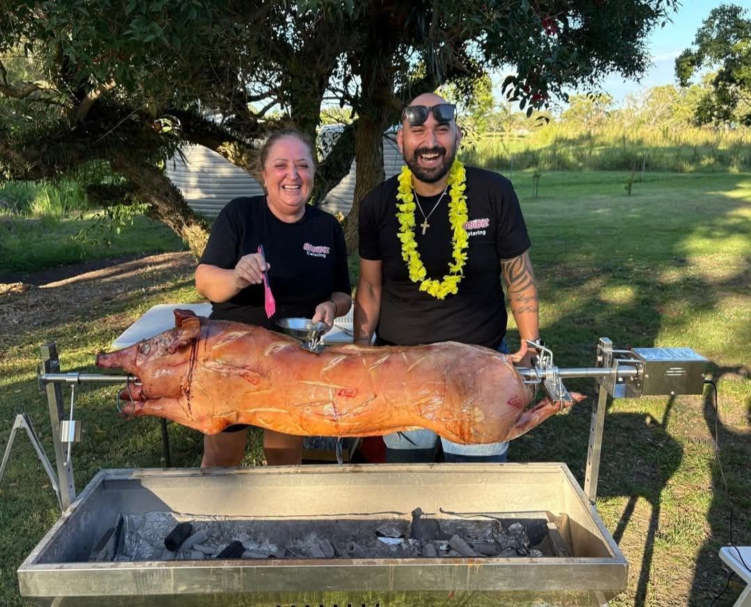 Two people smile, posing with roasted pig on a spit over a charcoal grill outdoors. — The Habibiz Catering In Wingham, NSW