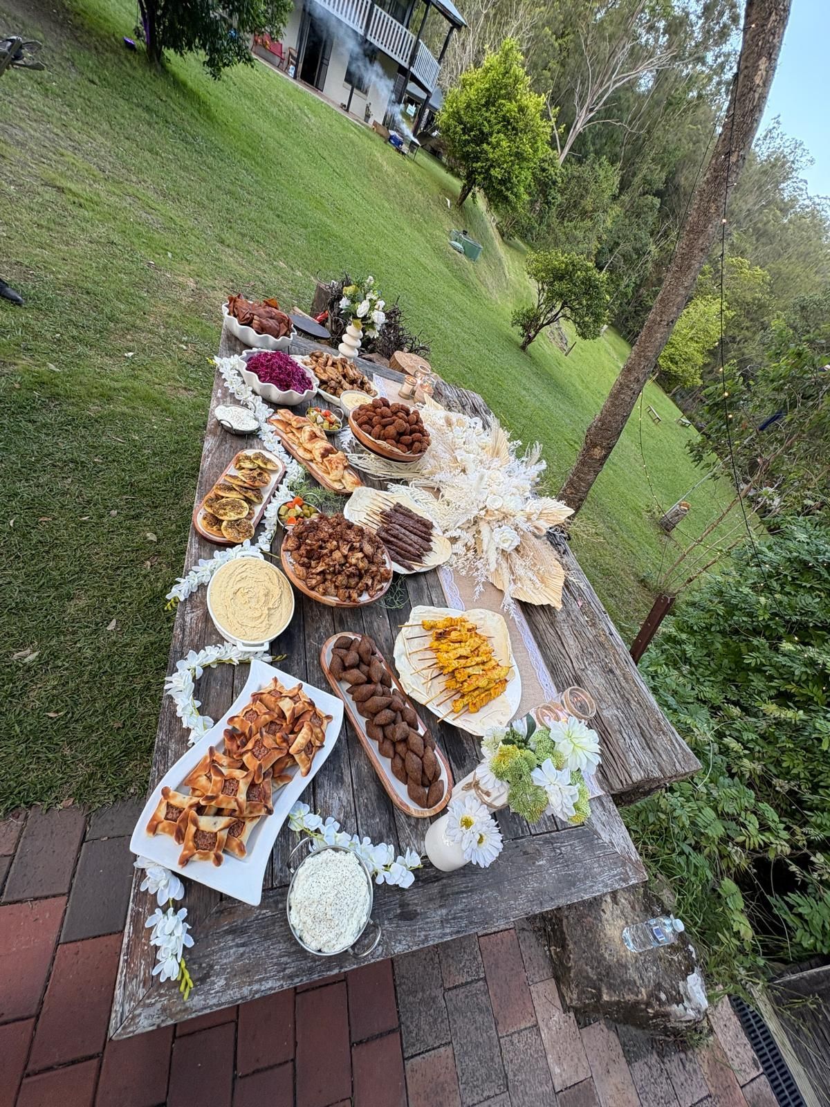 Wooden table laden with food, set outdoors on a grassy slope with a building in the background. — The Habibiz Catering In Wingham, NSW