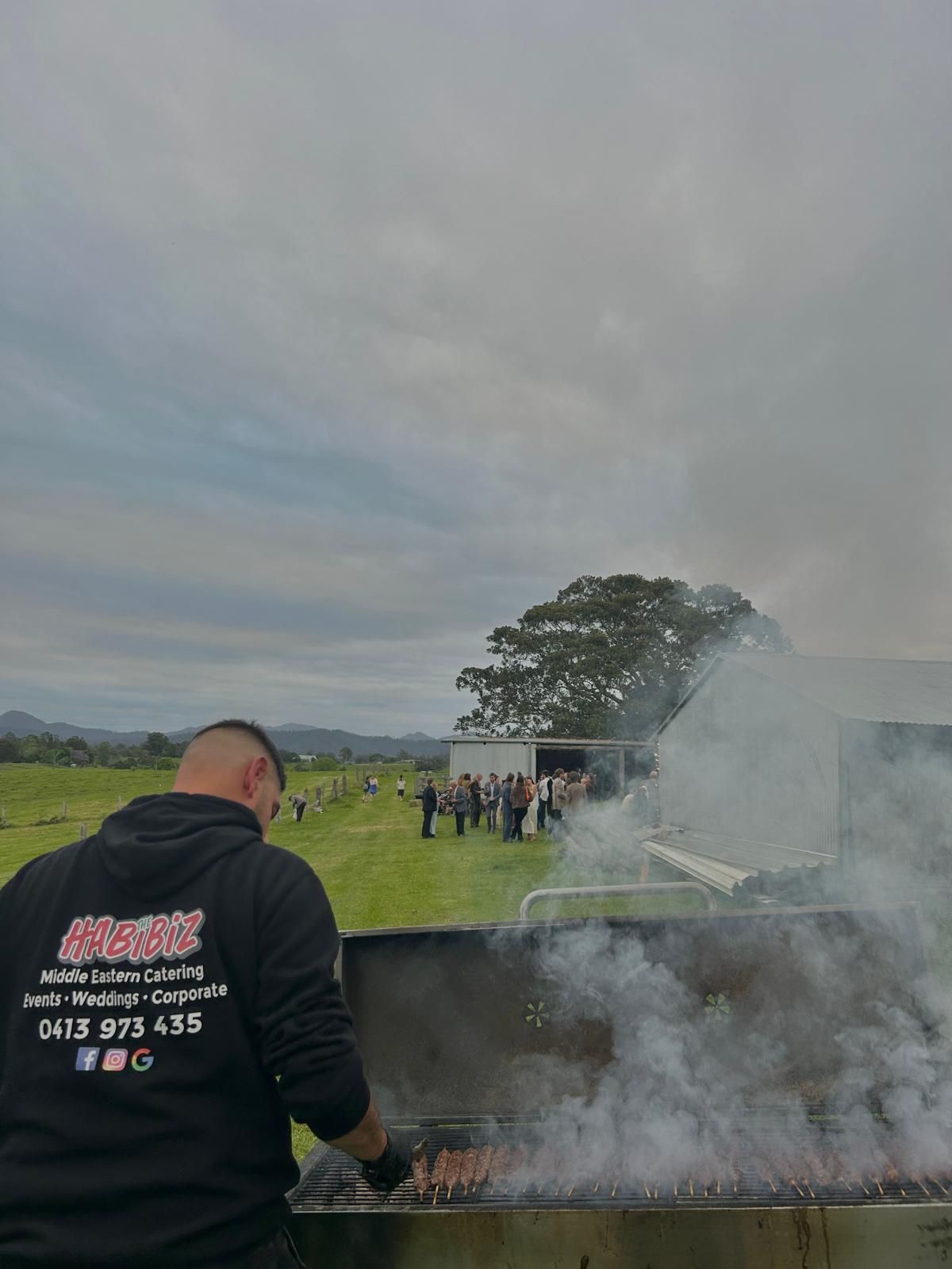 Man grilling outside, smoke rising. Cloudy sky, people in distance near building and field. — The Habibiz Catering In Wingham, NSW