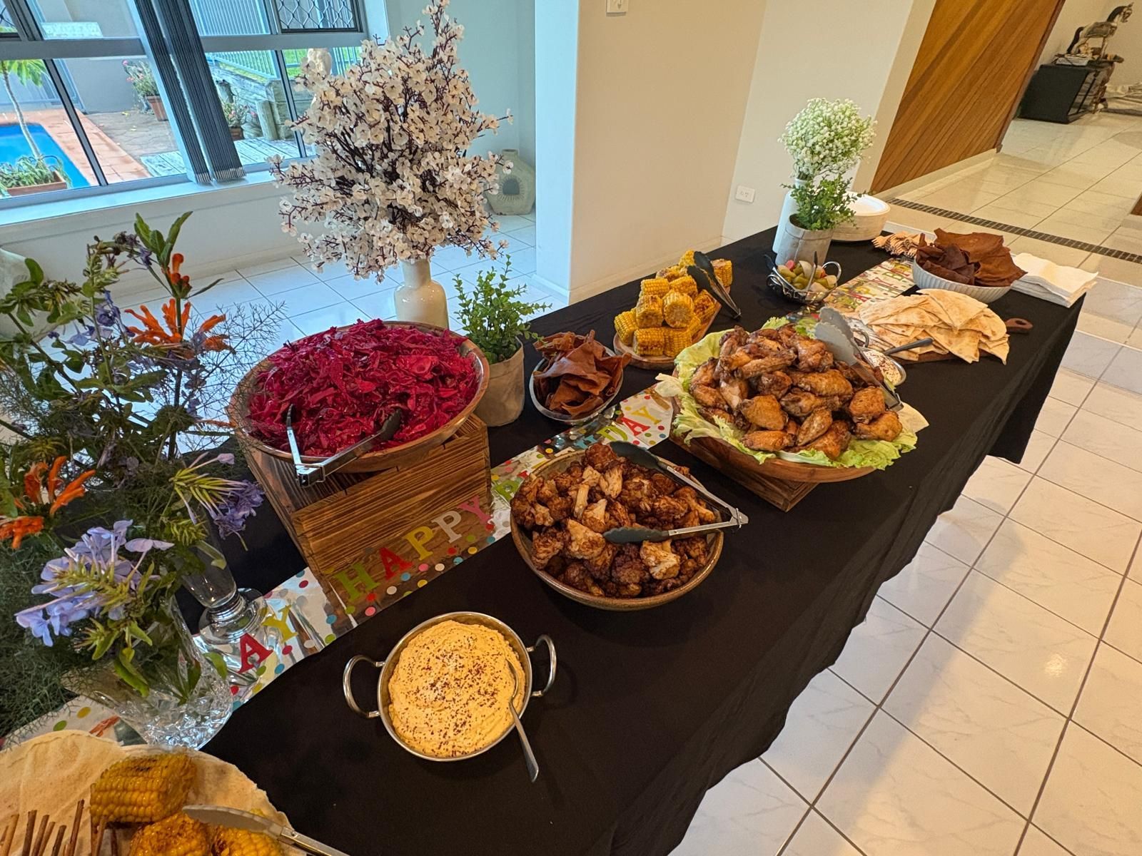 Buffet table with various dishes: chicken, potatoes, salad, pita bread, and dip. Decorated with flowers. — The Habibiz Catering In Wingham, NSW