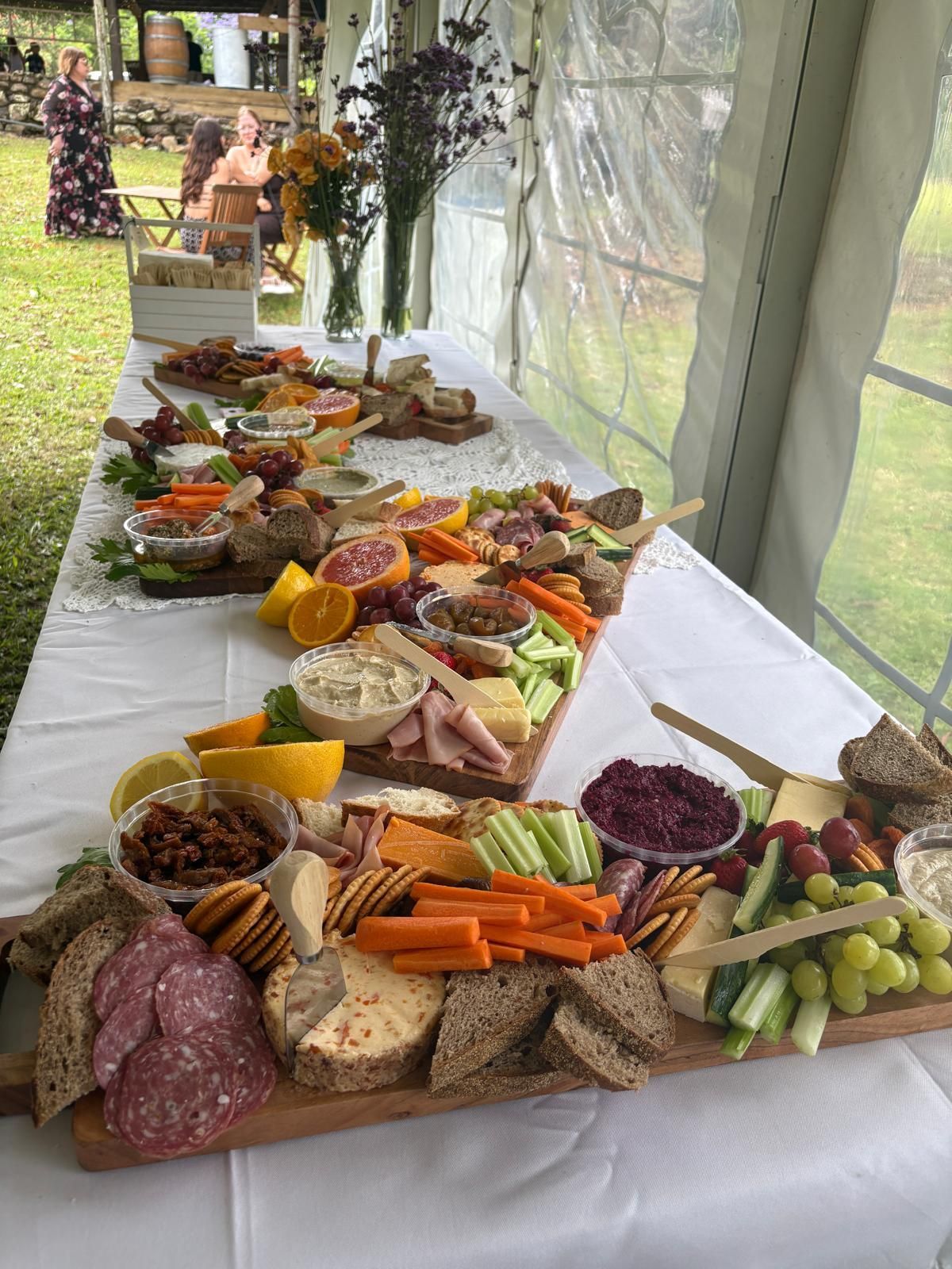 Long table laden with food, including cheeses, meats, crackers, vegetables, and dips; outdoors with people in the background. — The Habibiz Catering In Wingham, NSW