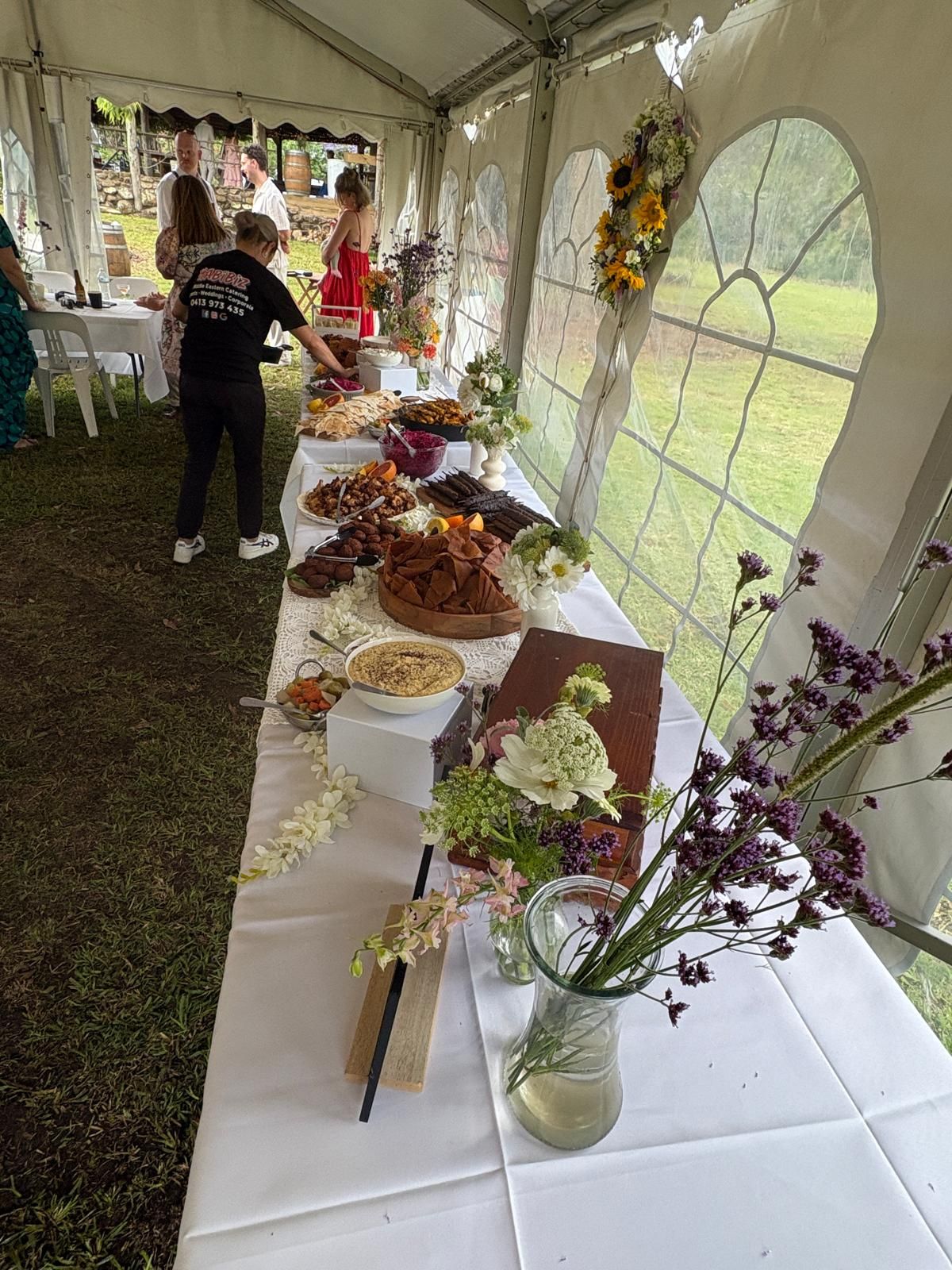 Food buffet table in a tent, with various dishes, flowers, and a person serving. — The Habibiz Catering In Wingham, NSW
