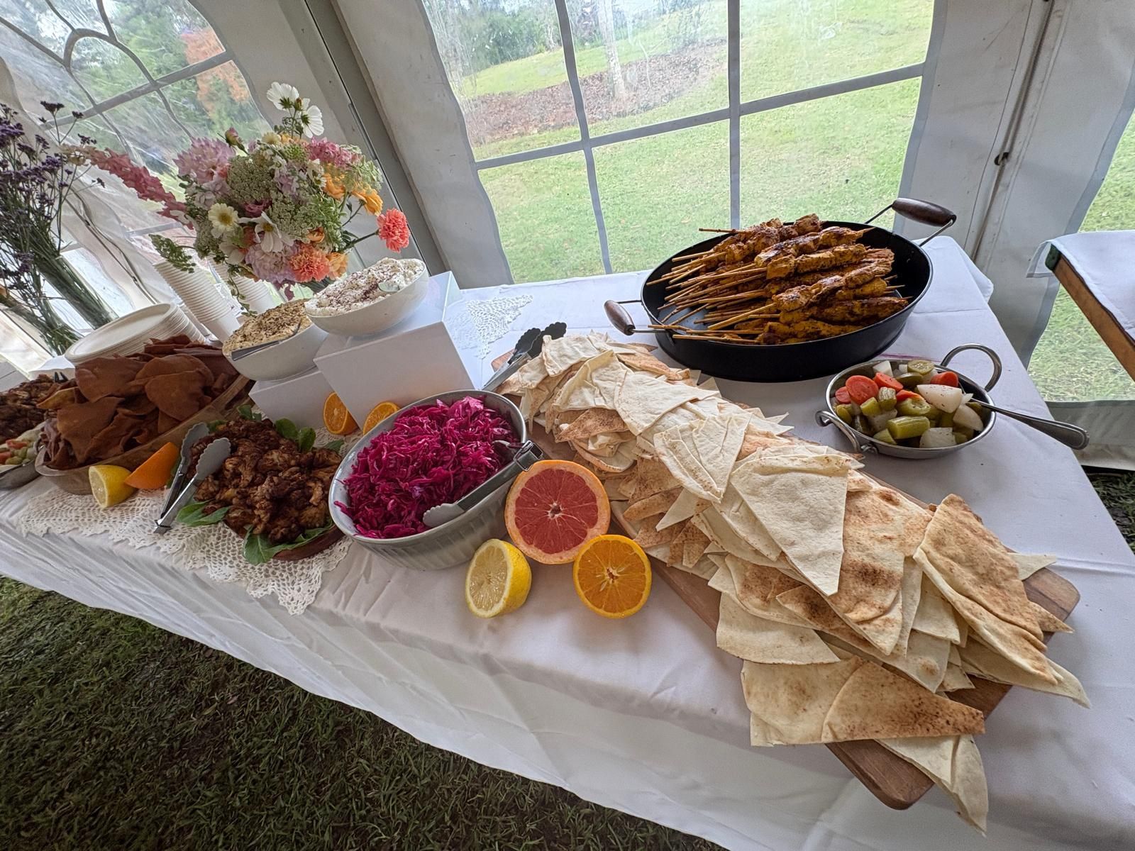 Food buffet on a table with a white cloth, inside a tent.  Includes bread, meat skewers, dips, fruit and flowers. — The Habibiz Catering In Wingham, NSW