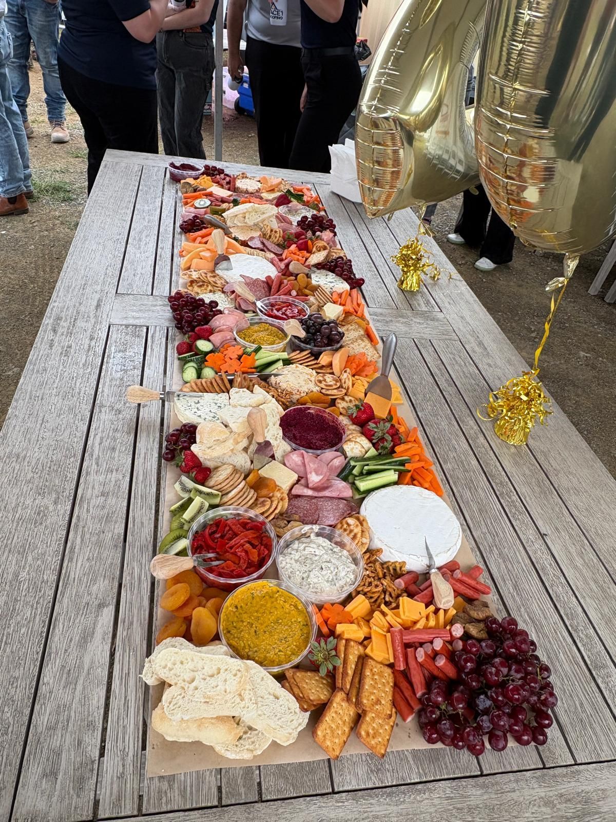 Long wooden table laden with charcuterie: cheeses, meats, crackers, dips, fruit, and vegetables. People stand nearby. — The Habibiz Catering In Wingham, NSW