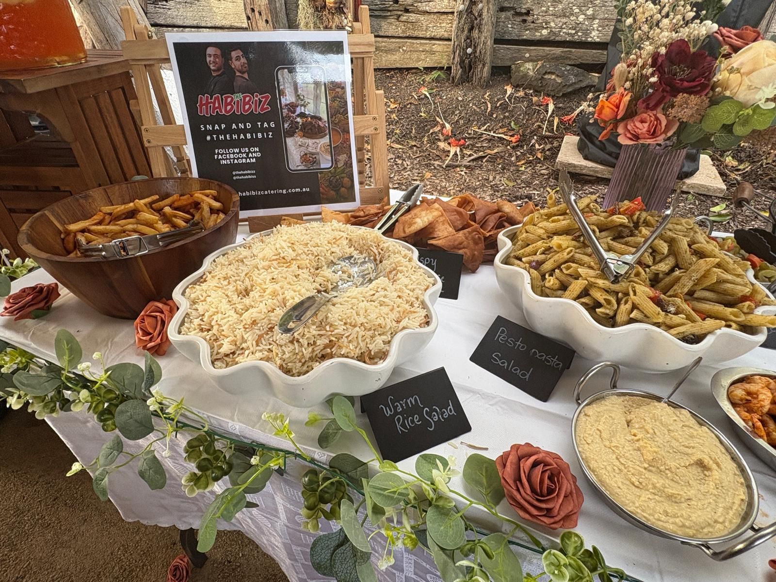 Food buffet with various dishes: rice, pasta, hummus, and fried items. Decorated with roses and greenery. — The Habibiz Catering In Wingham, NSW