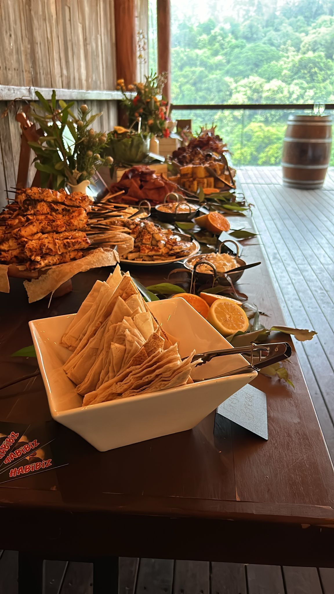A buffet table laden with food, next to a deck with a scenic view, flowers and a barrel. — The Habibiz Catering In Wingham, NSW