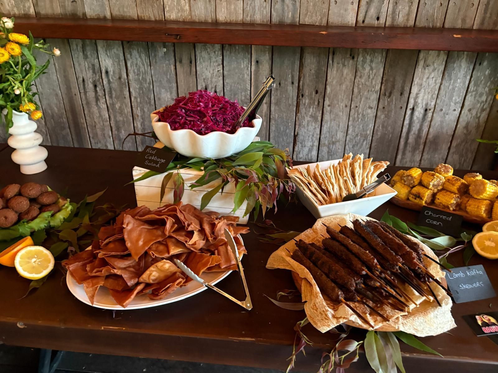Food buffet with kabobs, crackers, dips, and cookies on a wooden table, against a wooden wall. — The Habibiz Catering In Wingham, NSW
