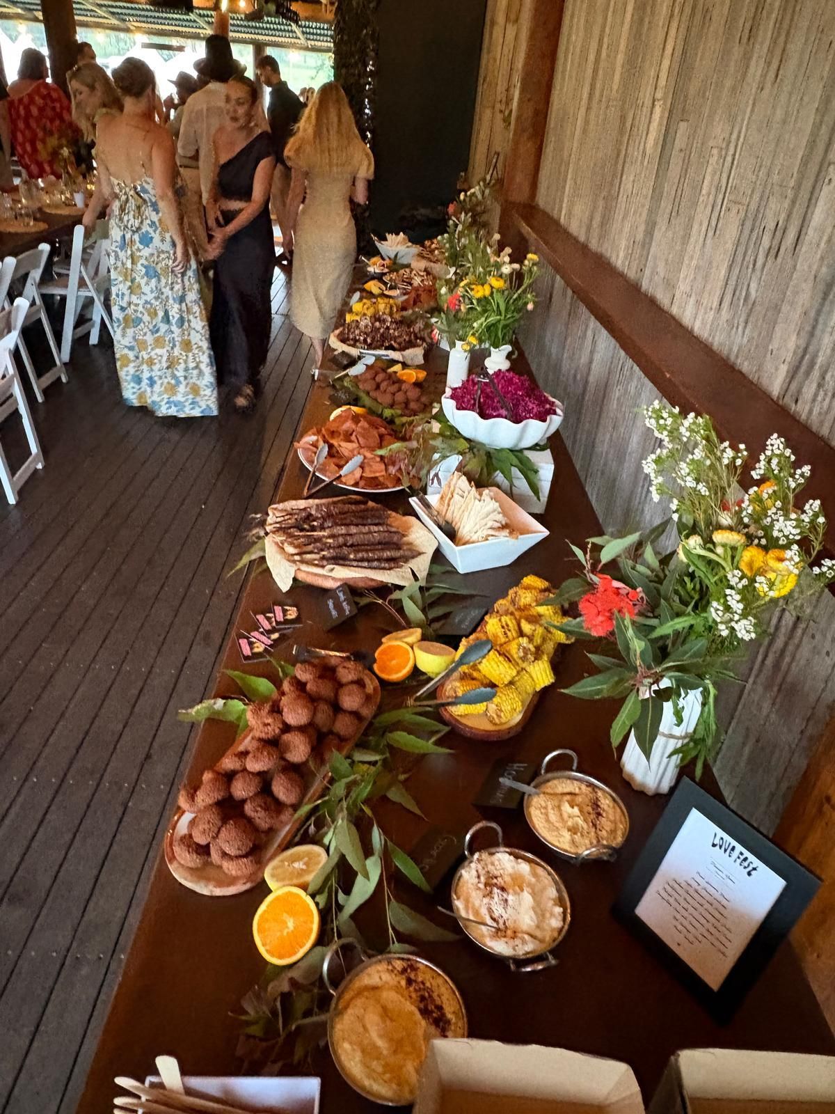 Food buffet table with various dishes and floral arrangements, inside a wood-paneled building; guests milling about. — The Habibiz Catering In Wingham, NSW