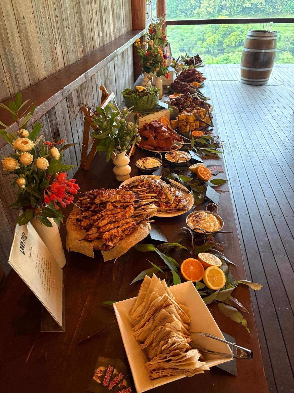 Buffet table with assorted food and flowers, wooden setting, window view. — The Habibiz Catering In Wingham, NSW