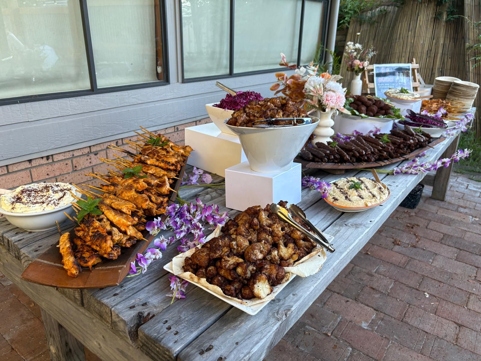 A buffet table with various dishes: grilled skewers, cauliflower, and dips, decorated with flowers. — The Habibiz Catering In Wingham, NSW