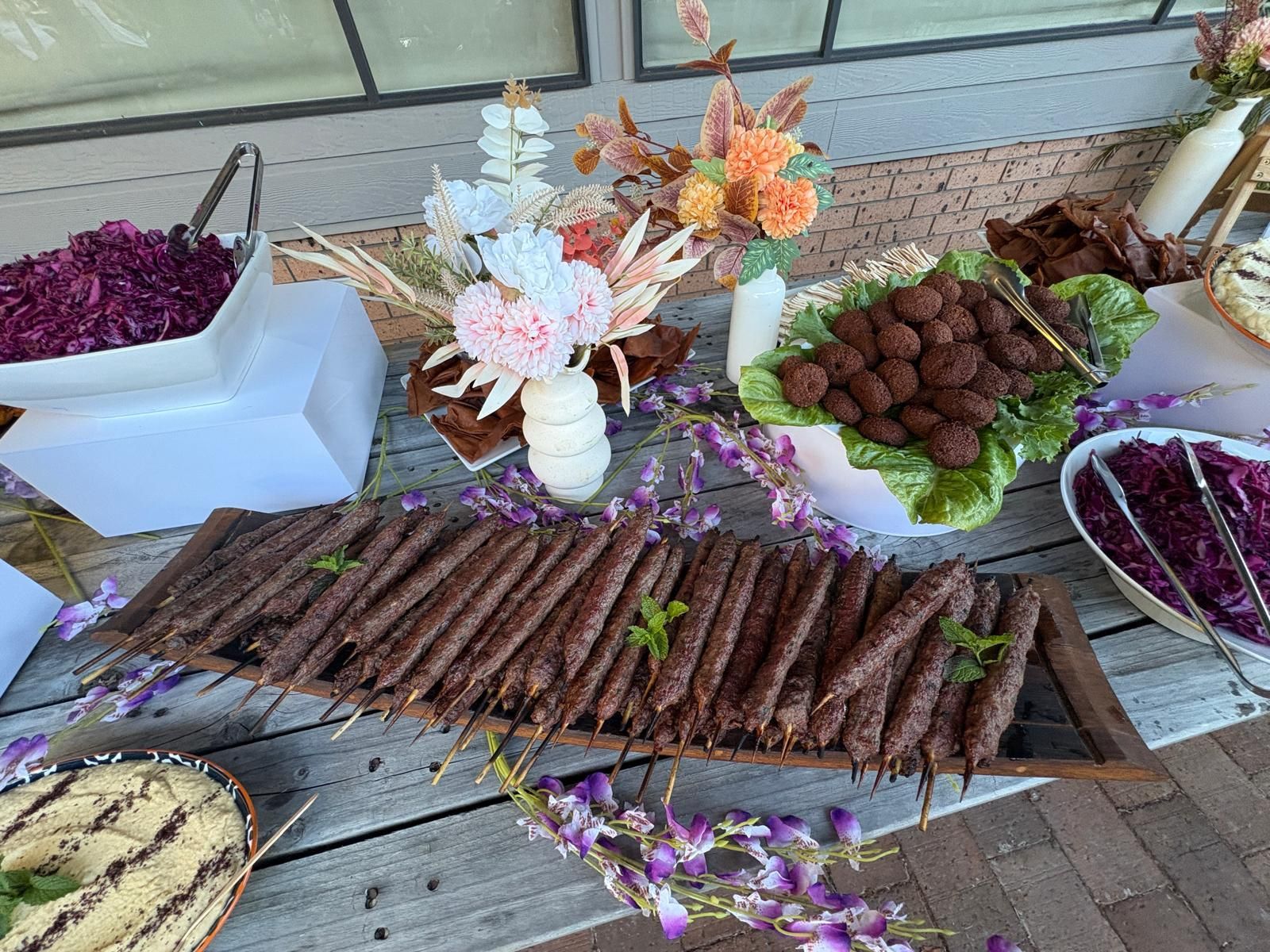 Buffet table with various dishes: kebabs, meatballs, salads, and hummus, decorated with flowers. — The Habibiz Catering In Wingham, NSW