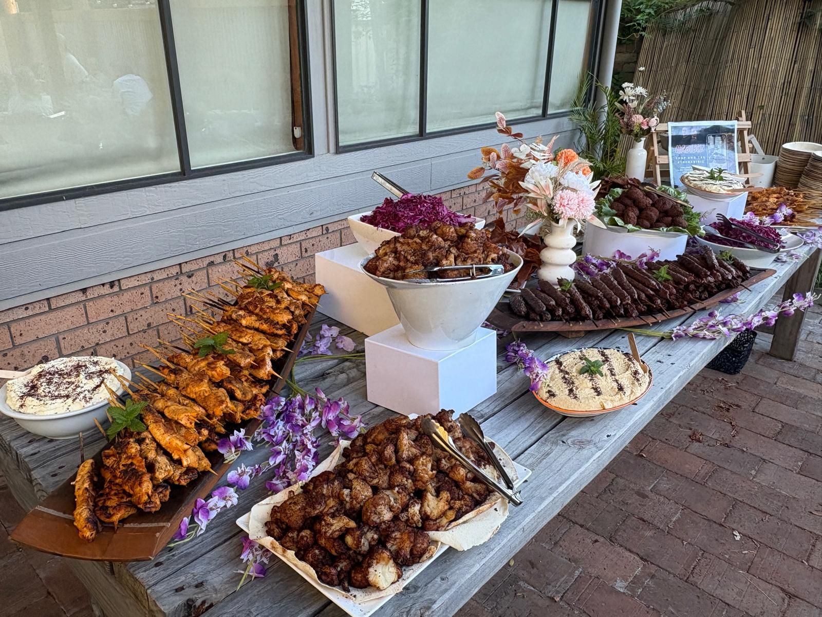 Food buffet with various dishes and floral decorations on a wooden table outdoors. — The Habibiz Catering In Wingham, NSW