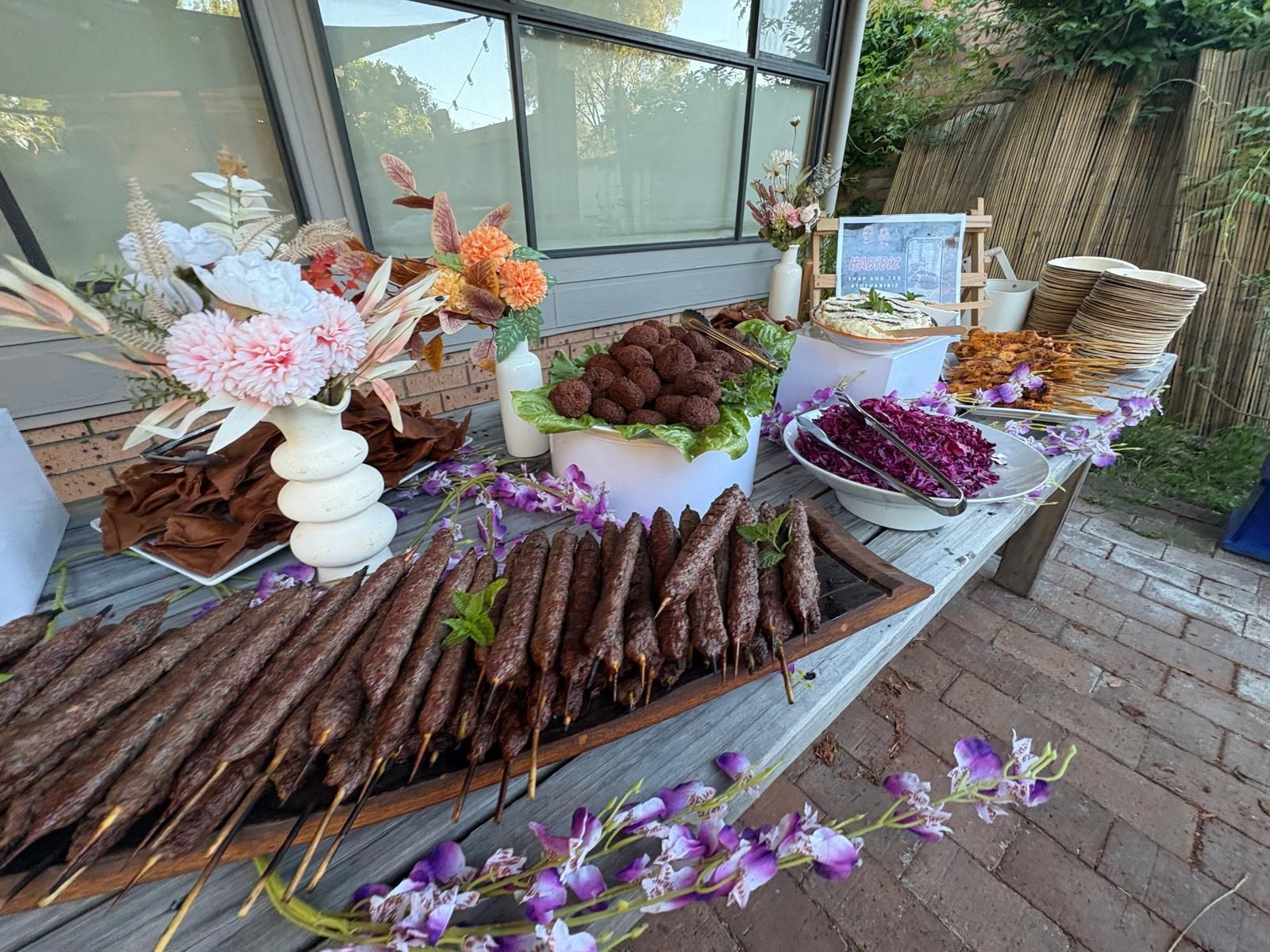 Food spread on a wooden table: kebabs, falafel, dips, and floral decorations. Outdoor setting with greenery and brick. - The Habibiz Catering in Wingham NSW